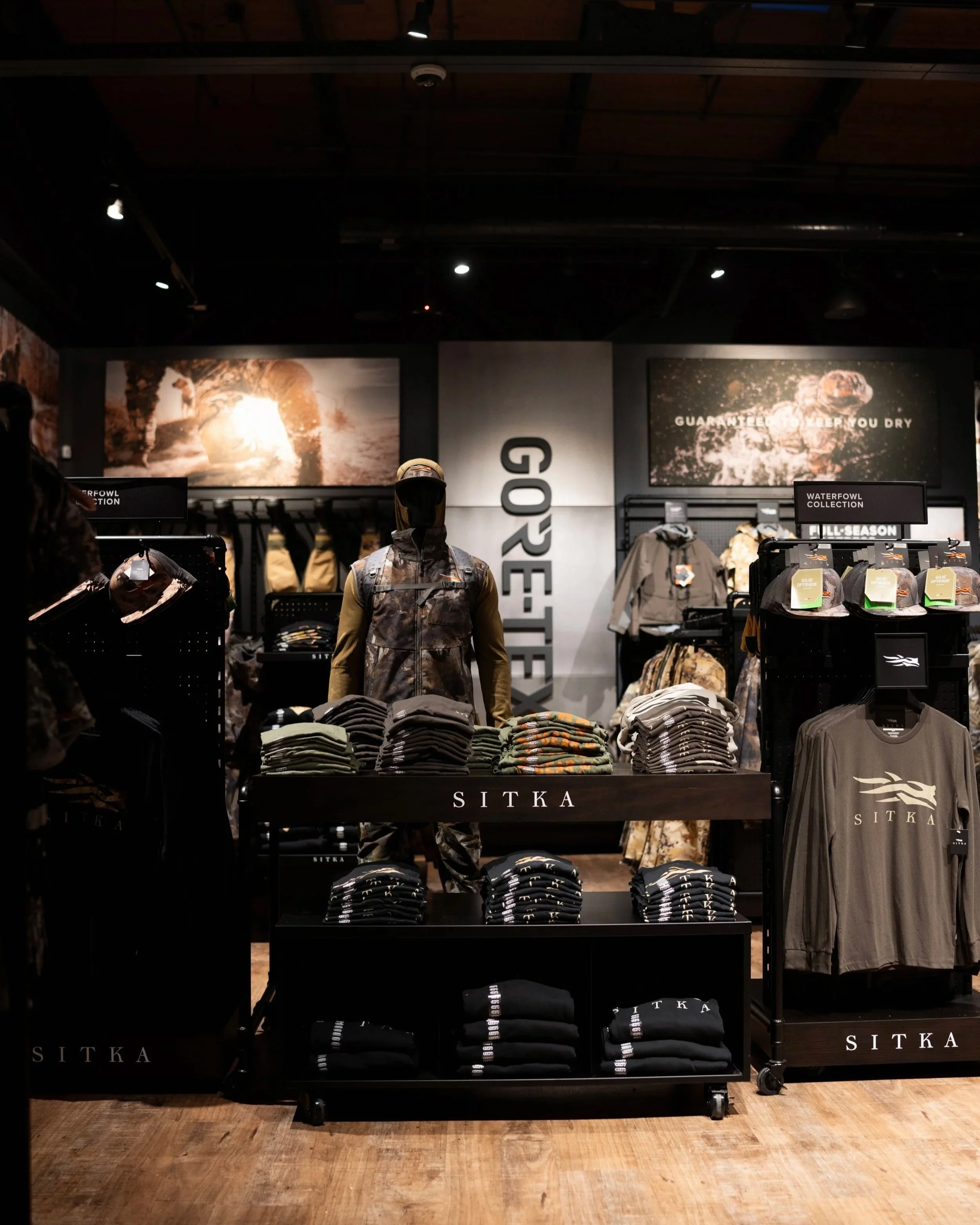 A clothing display in a store featuring men's outdoor apparel, including camouflage jackets and hats, with promotional posters and signage in the background.