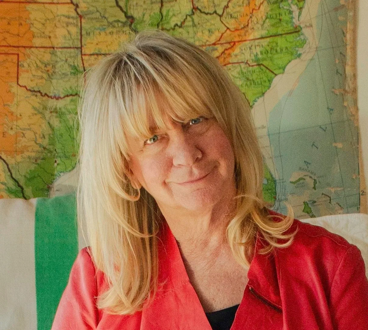 A smiling woman with blonde hair and bangs, wearing a red jacket, sitting in front of a colorful map of the southeastern United States.