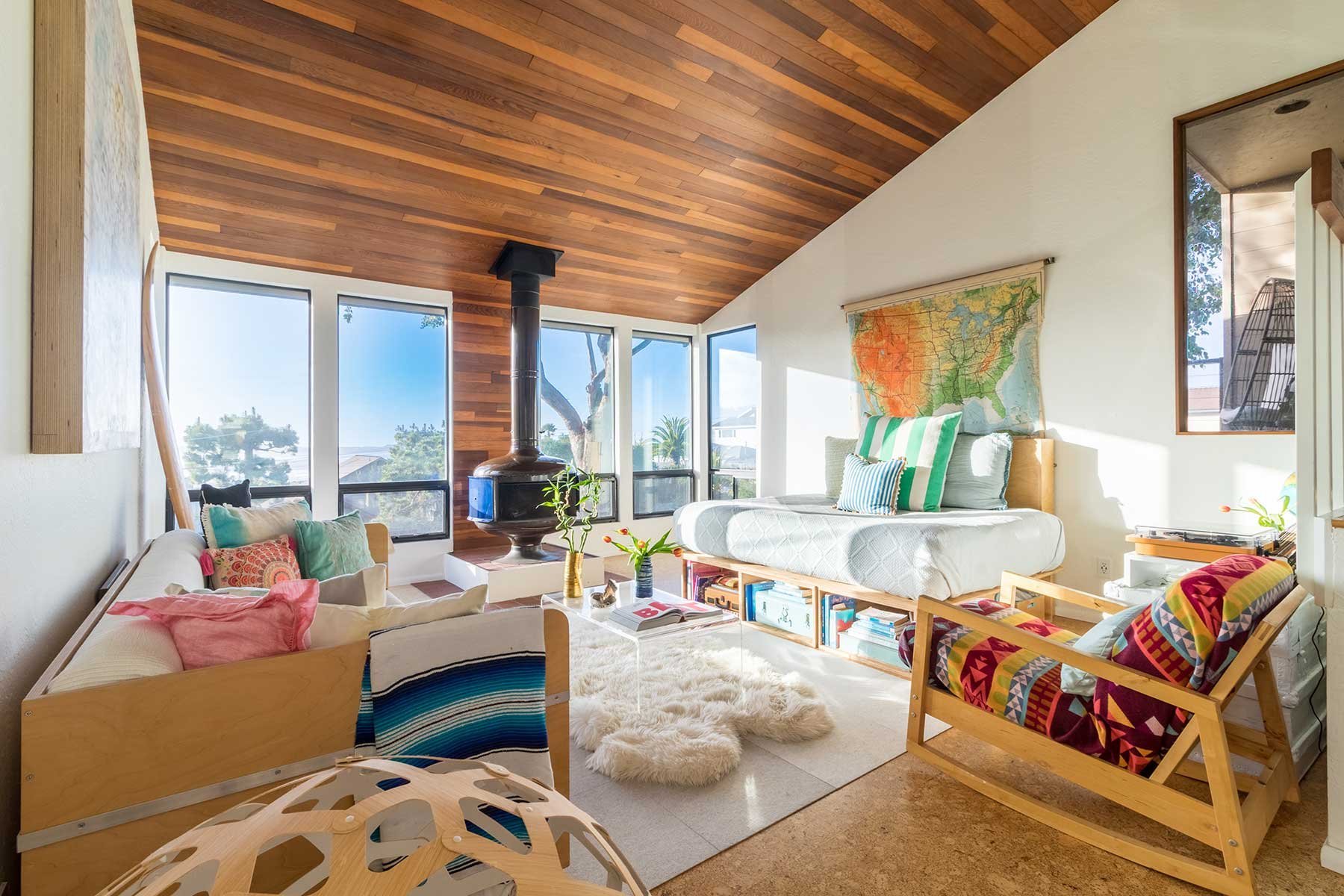 Living room with wooden ceiling, large windows, and a wood-burning stove. Contains a white sofa with colorful pillows, a daybed, a armchair with a vibrant patterned cover, a coffee table, and various plants and books, with a map of the United States on the wall.