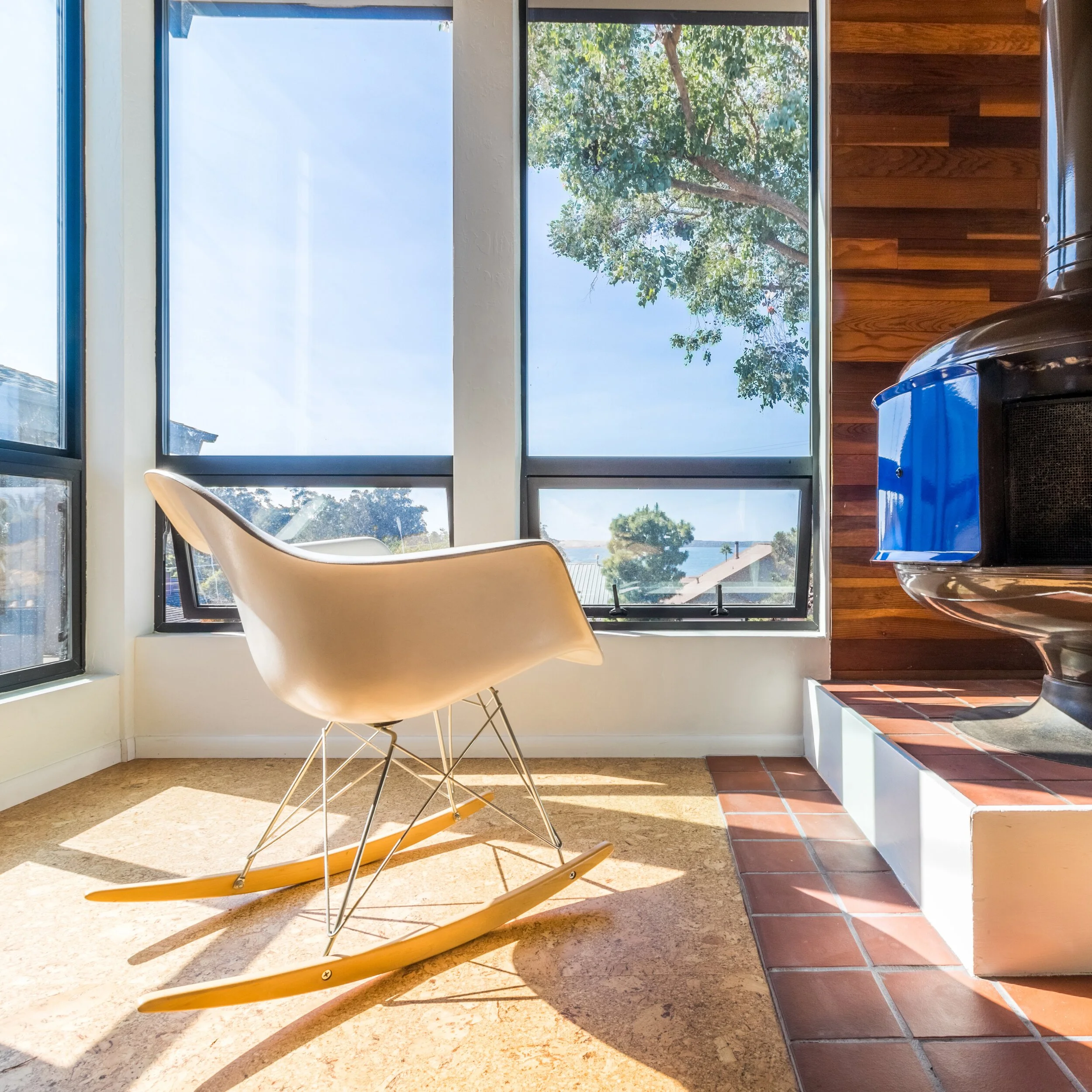 A rocking chair on a beige carpet near a large window with a view of trees and the ocean in the distance, a blue container on a wooden countertop, and a black and silver kitchen appliance.