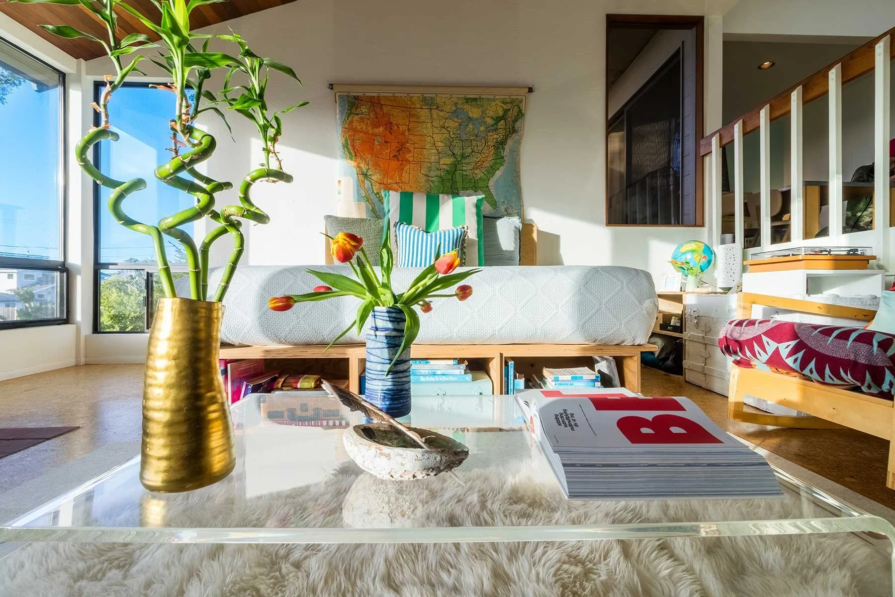 Living room with sunlight, featuring potted plants in gold and blue vases on glass coffee table, a bed with striped pillows, a map on the wall, books on a wood shelf, and a plush white rug.