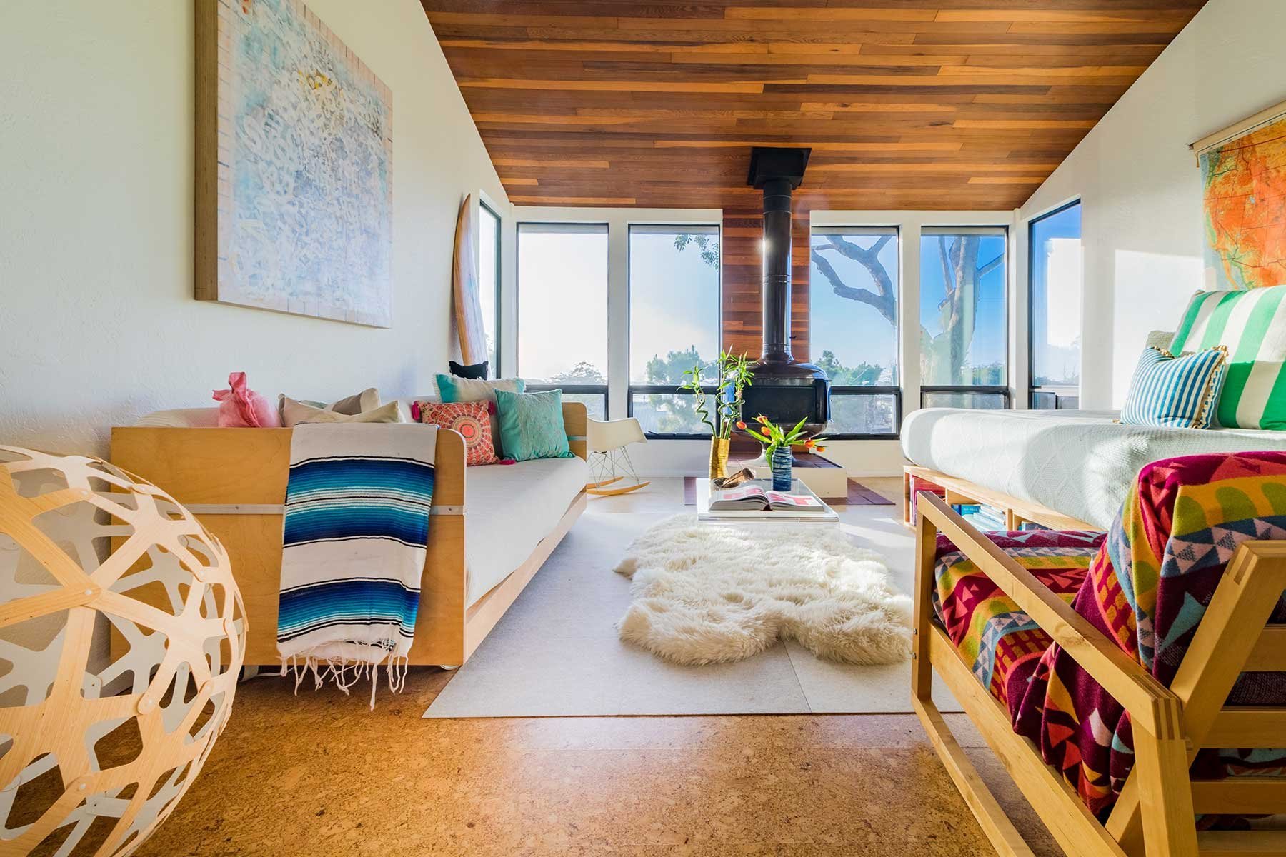 A bright living room with wooden ceiling and large windows, decorated with multicolored pillows, plants, and artwork. There's a cozy white rug and a wood stove in the corner.