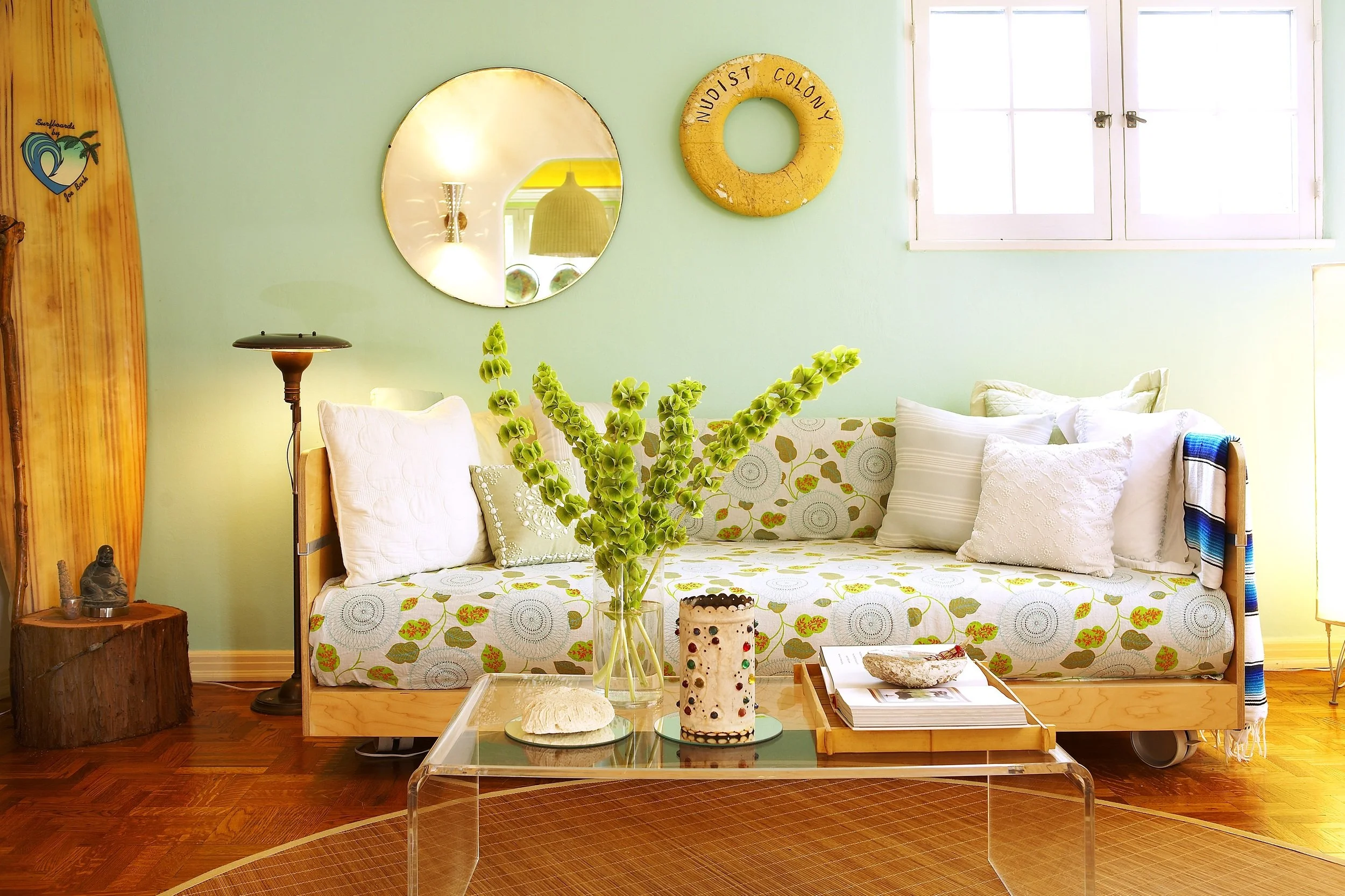 Living room with green walls, a floral-patterned sofa with white pillows, a glass coffee table with decorative candles, a tall green plant in a glass vase, a mirror and a yellow lifebuoy on the wall, and windows allowing natural light.
