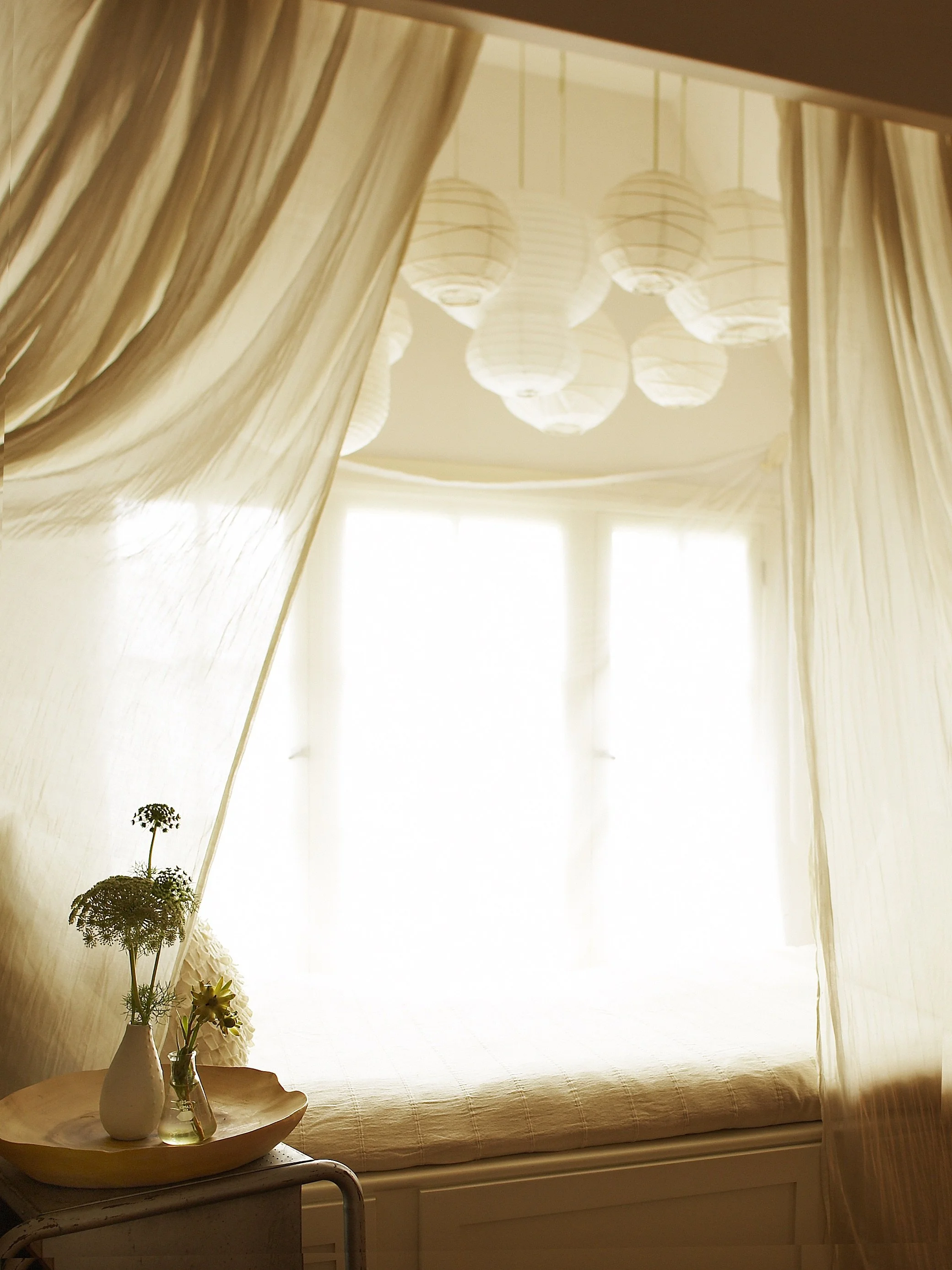 The image shows a cozy corner with soft beige curtains framing a window with sheer white drapes, illuminating a simple bed with a beige bedspread. A small side table holds a white pitcher and a glass vase with flowers.