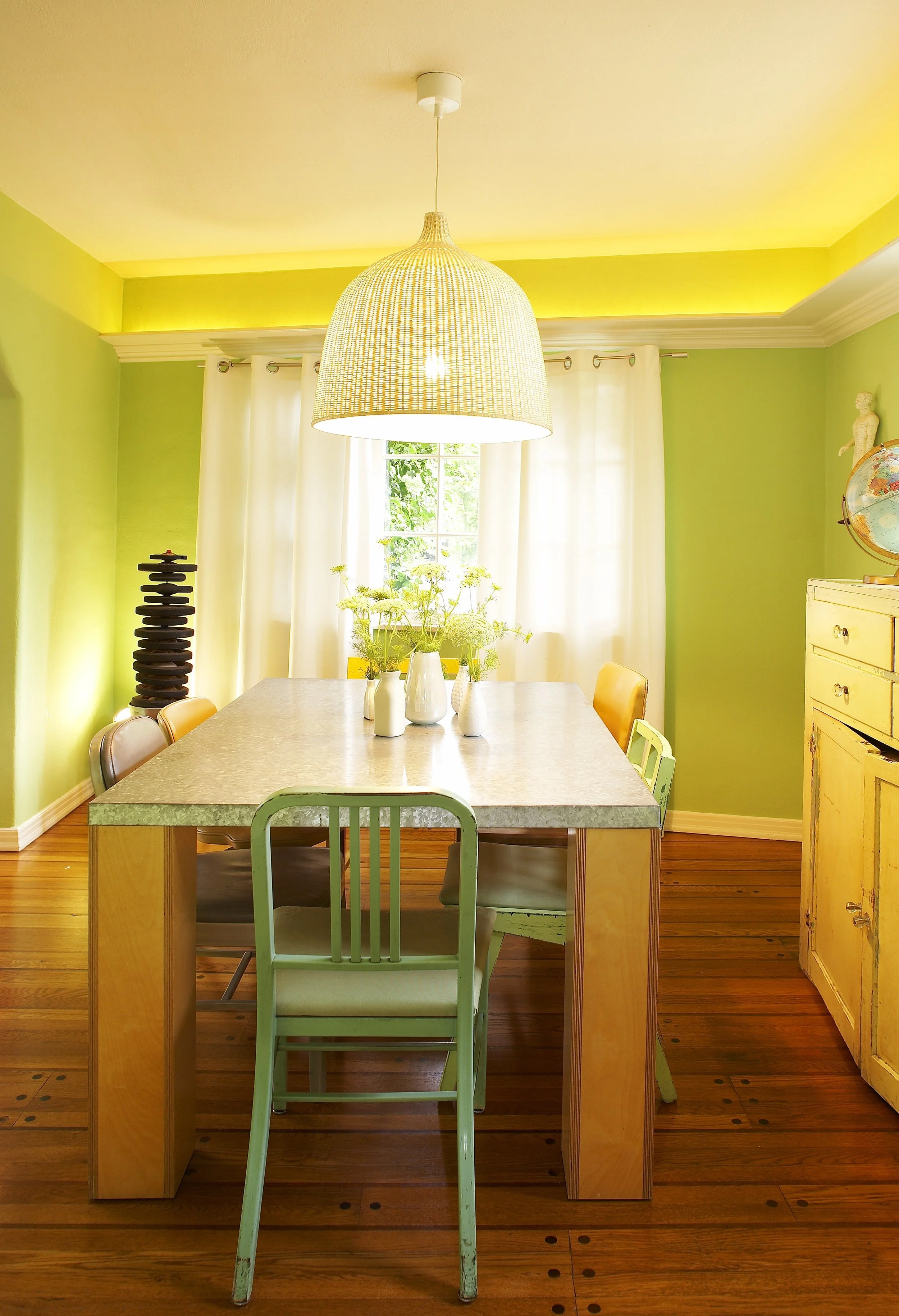 A dining room with a large wooden table, surrounded by vintage chairs in pastel colors. A large woven pendant light hangs above the table. The room has lime green walls with white curtains on the window, a yellow ceiling accent, and a wooden floor. T