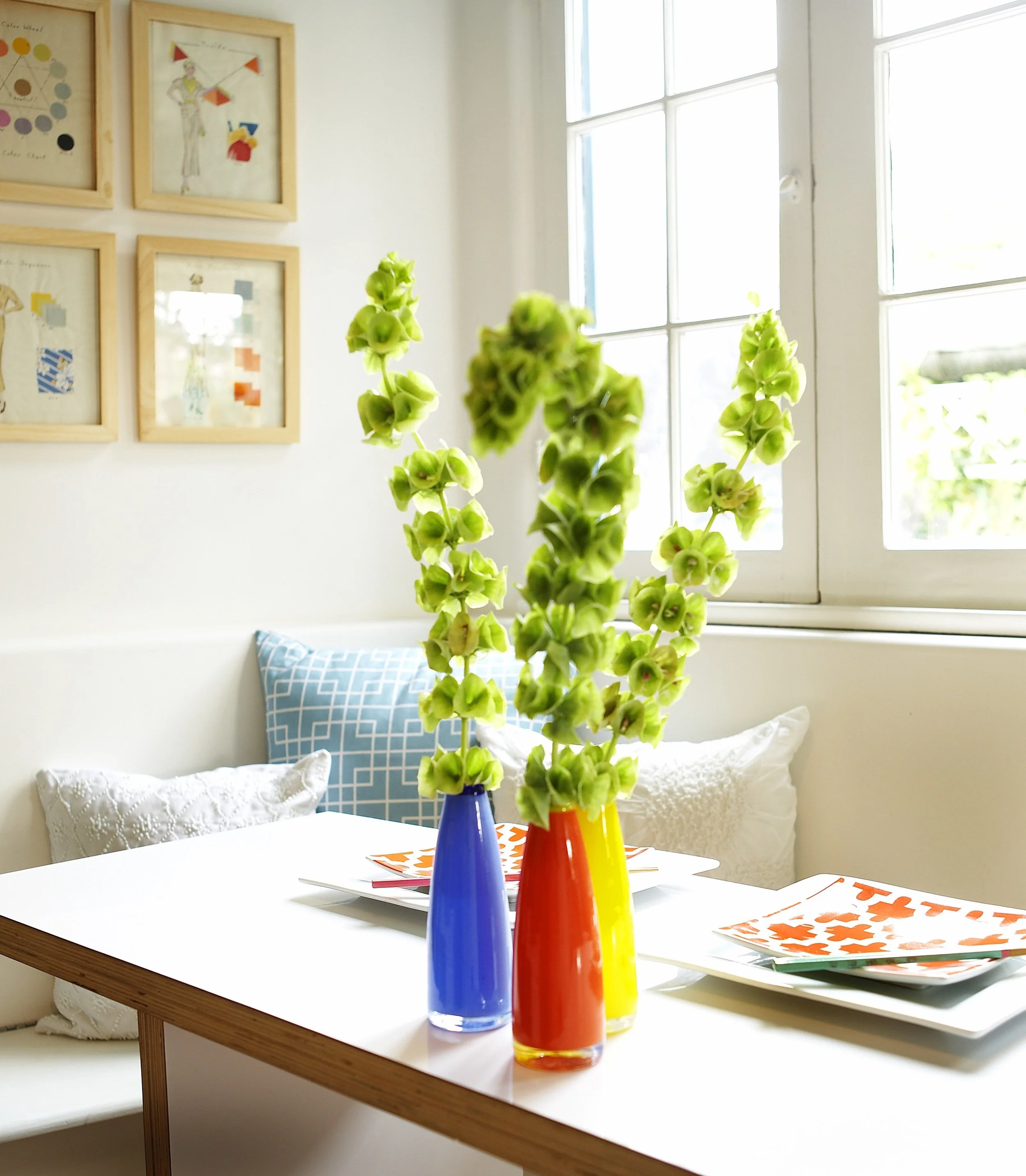 Vases with green flowers on a dining table near a window, with colorful paper or artwork on the table, and framed artwork on the wall in the background.