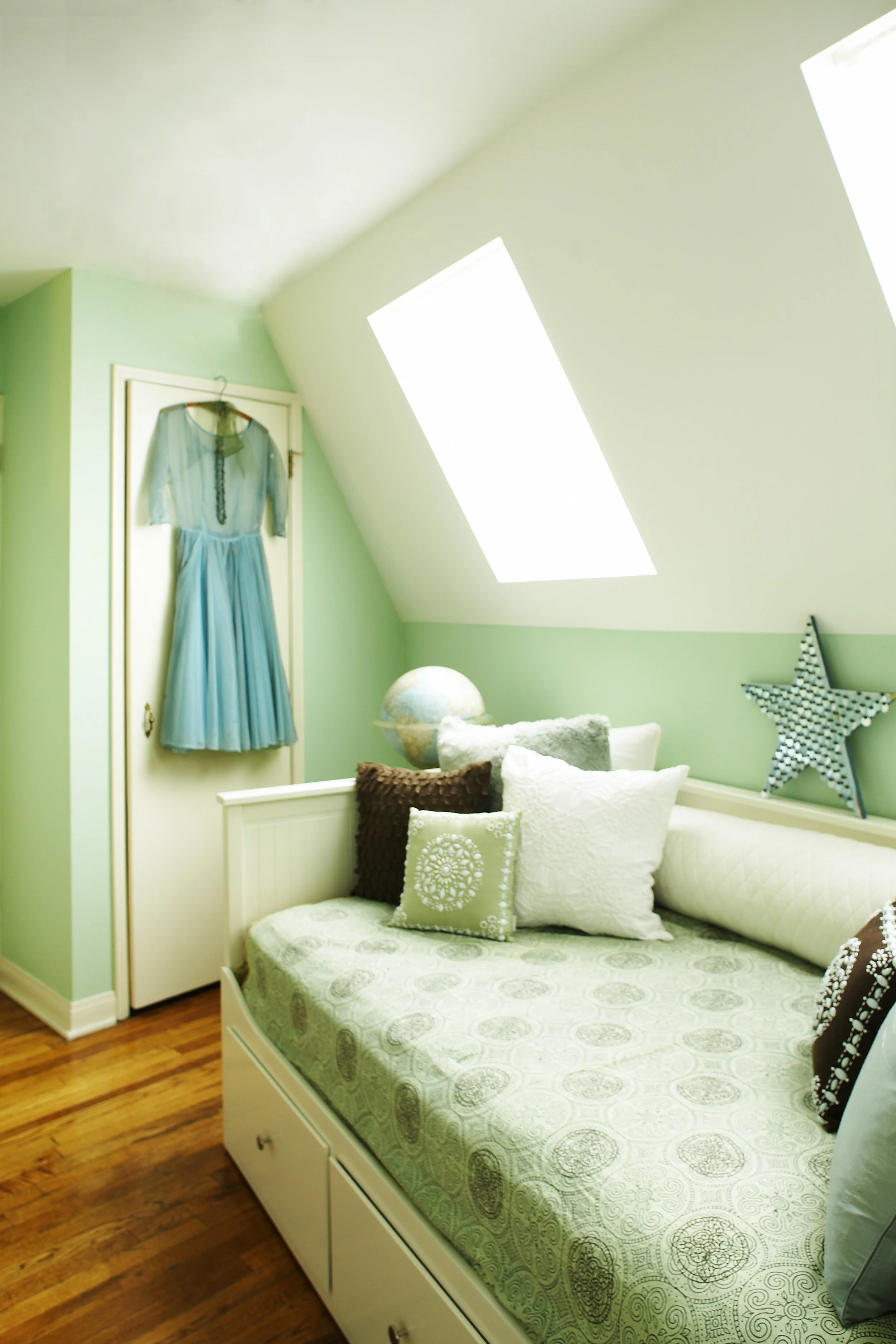 A cozy bedroom with a white bed, multiple pillows, and a green and white patterned bedspread. The wall behind the bed is painted light green, and there is a blue dress hanging on a white door. There is a globe on the bedside table, a star-shaped deco