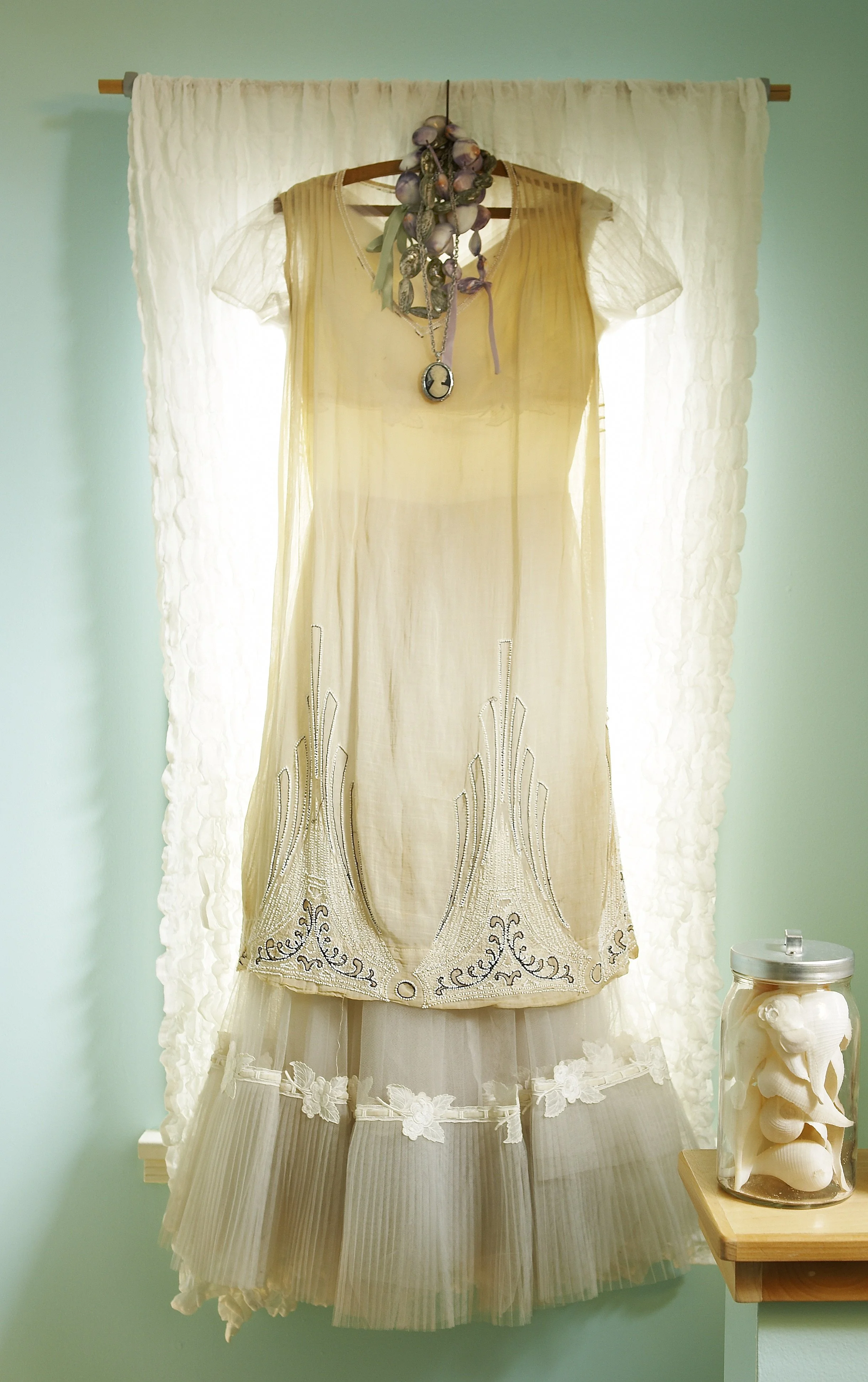 A vintage wedding dress with intricate embroidery, lace, and pleats hanging on a wooden rod against a light teal wall, with a floral jewelry necklace and a sheer overlay with floral patterns on a table beside it.