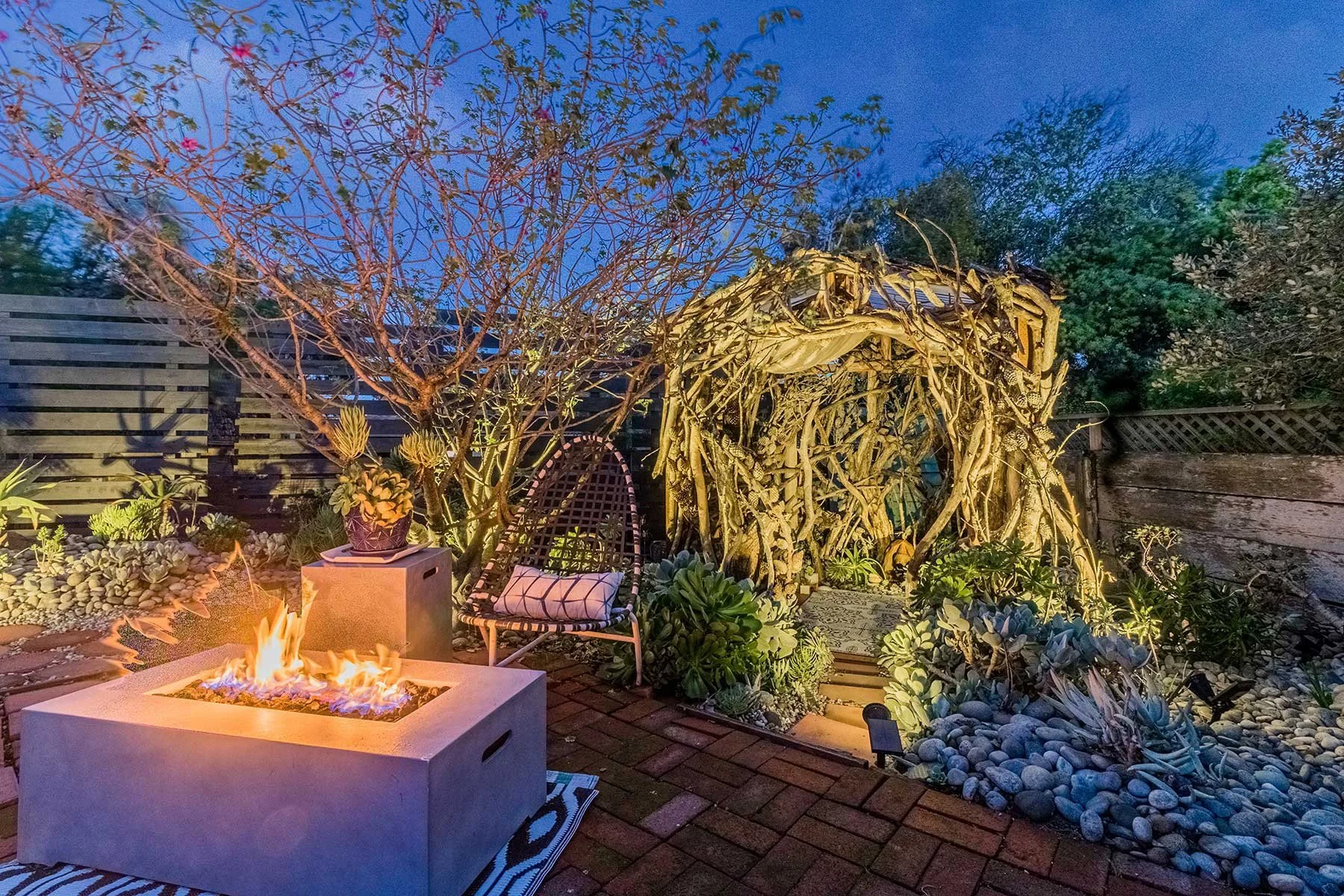 A backyard patio at dusk with a fire pit, a black chair with a white pillow, a wooden archway made of intertwined branches, and lush plants and succulents along a wooden fence and garden bed.