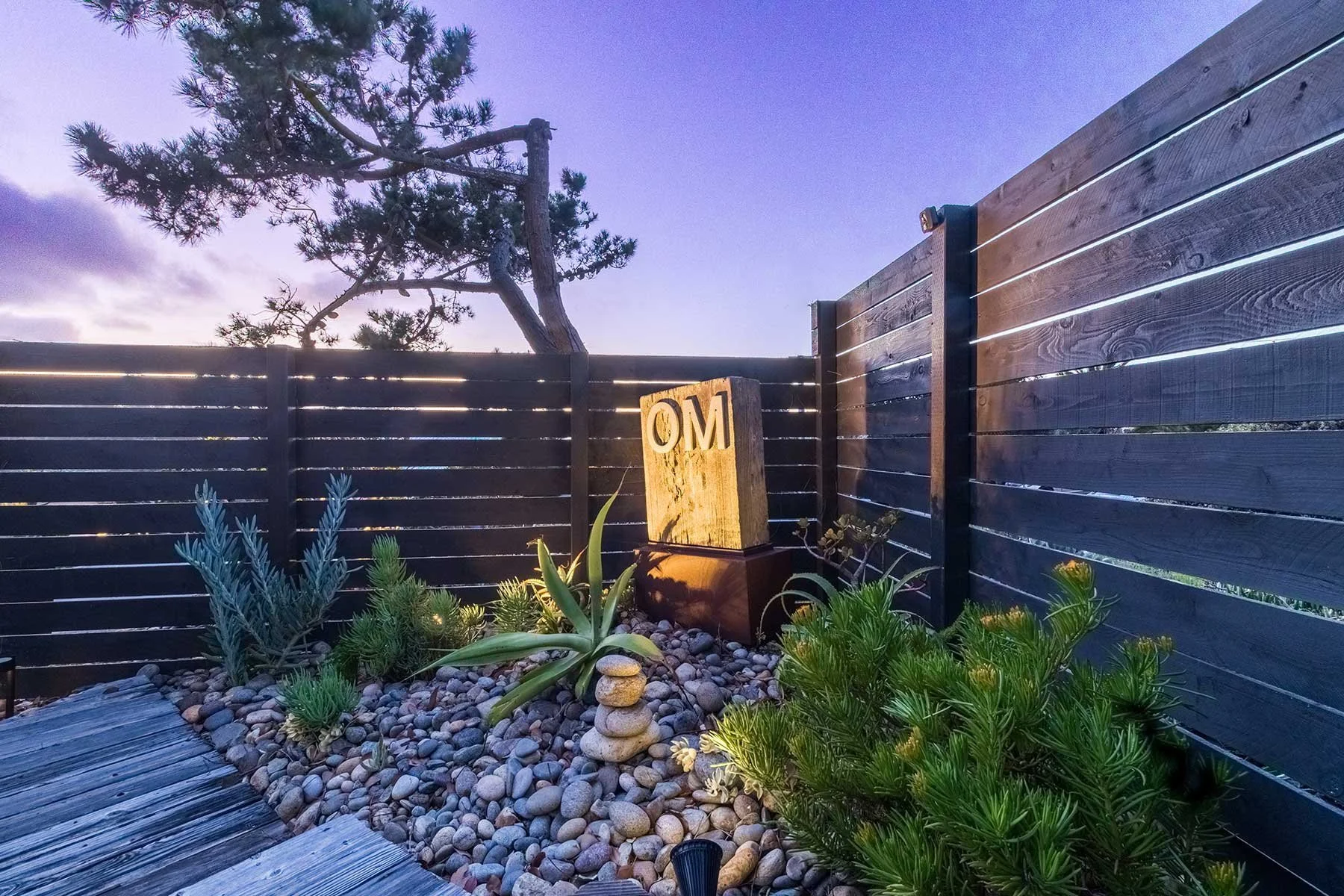 A small outdoor garden area enclosed by dark wooden fence panels, with a tree in the background, and illuminated by exterior lighting. There is a carved wooden block with the initials 'OM', surrounded by various green plants, succulents, and a small 