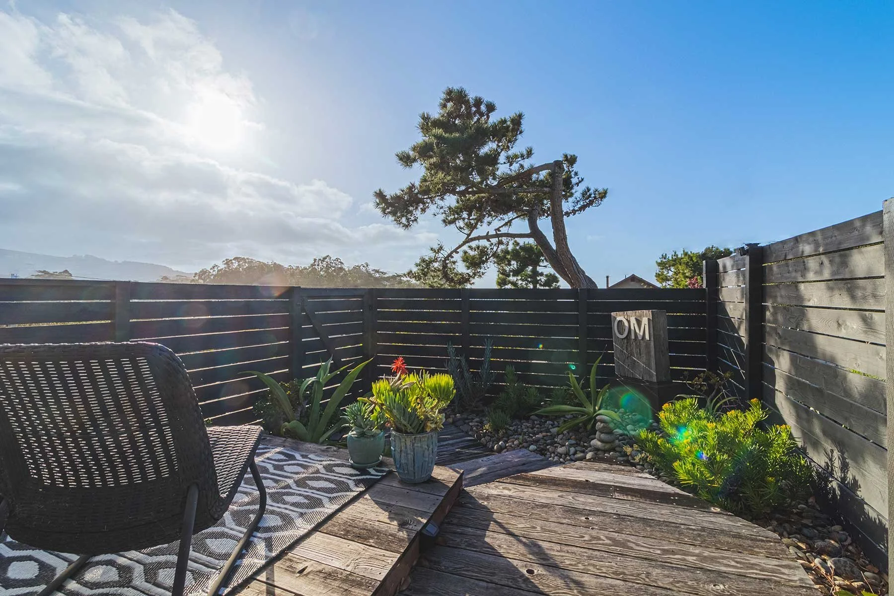 A backyard patio with wooden decking, wicker chair, potted succulents, and a black wooden fence. A large tree with bent branches and a bright sun overhead. The sky is partly cloudy with mountains in the background.