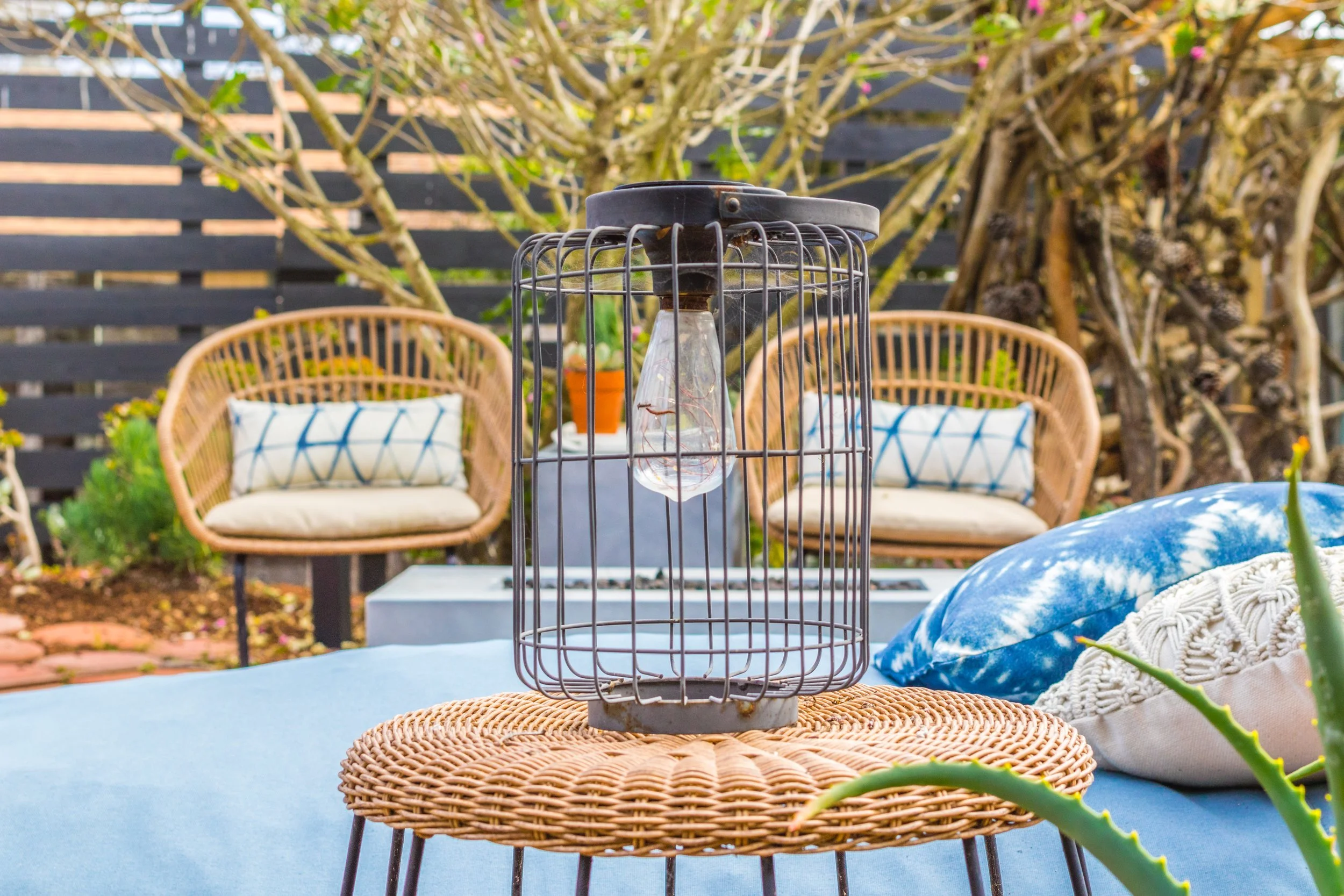 Outdoor patio with wicker chairs, a table, cushions, a garden fence, and a birdcage with a light bulb, surrounded by plants and trees.