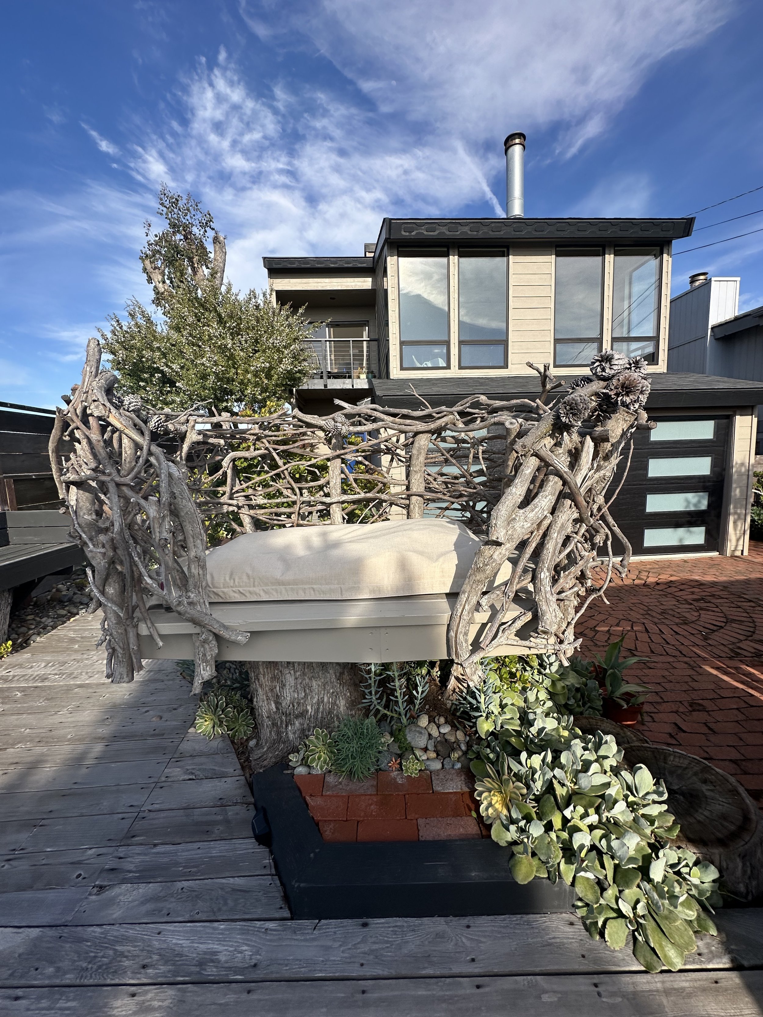 A wooden deck with hanging plants and a large wicker chair. In the background, there is a modern house with large windows, a balcony, and a garage. The sky is blue with some clouds.