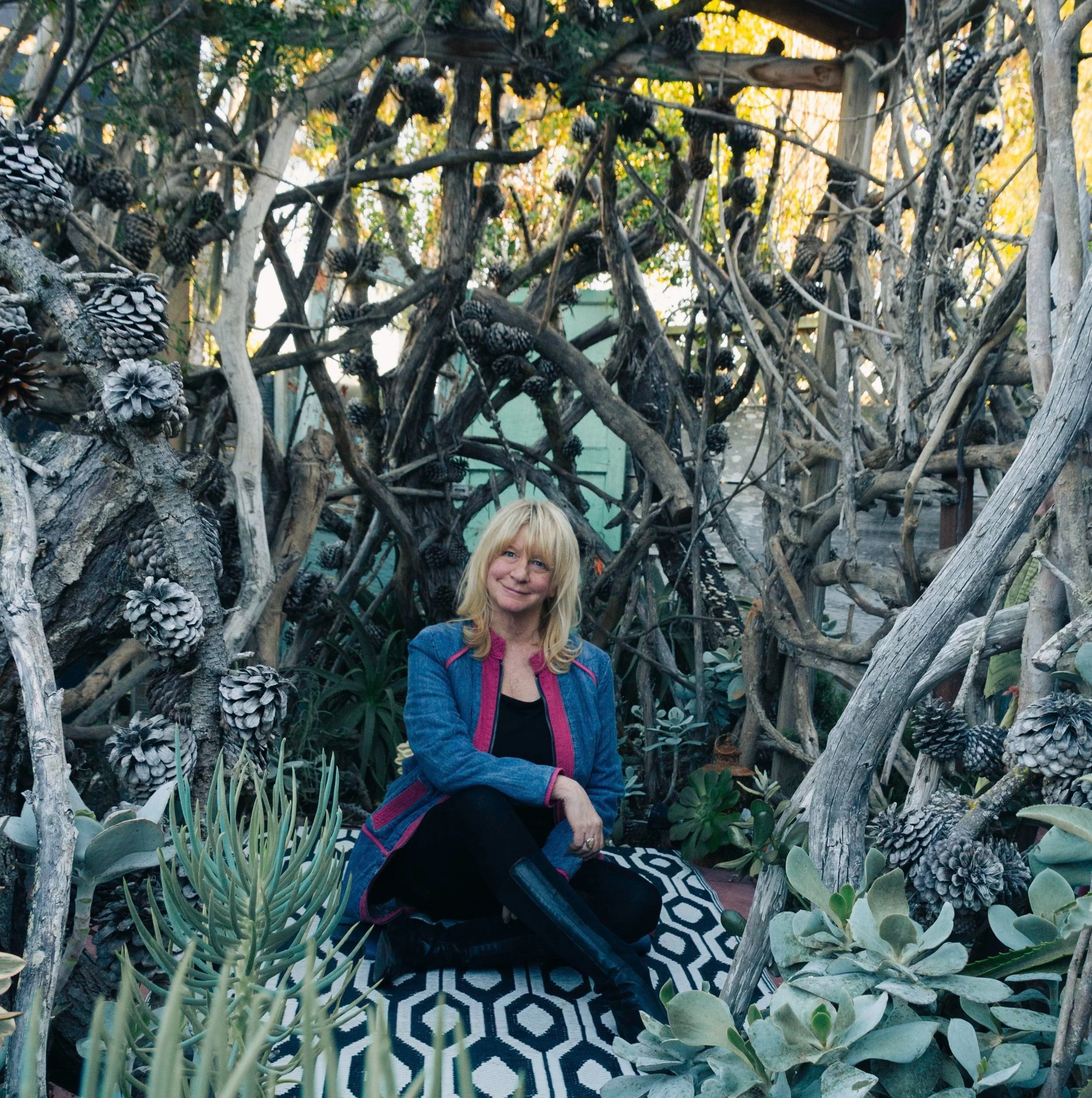 A woman with blonde hair, wearing a blue denim jacket and black pants, sitting on a patterned outdoor rug, surrounded by a dense arrangement of tree branches, pine cones, and various succulents.