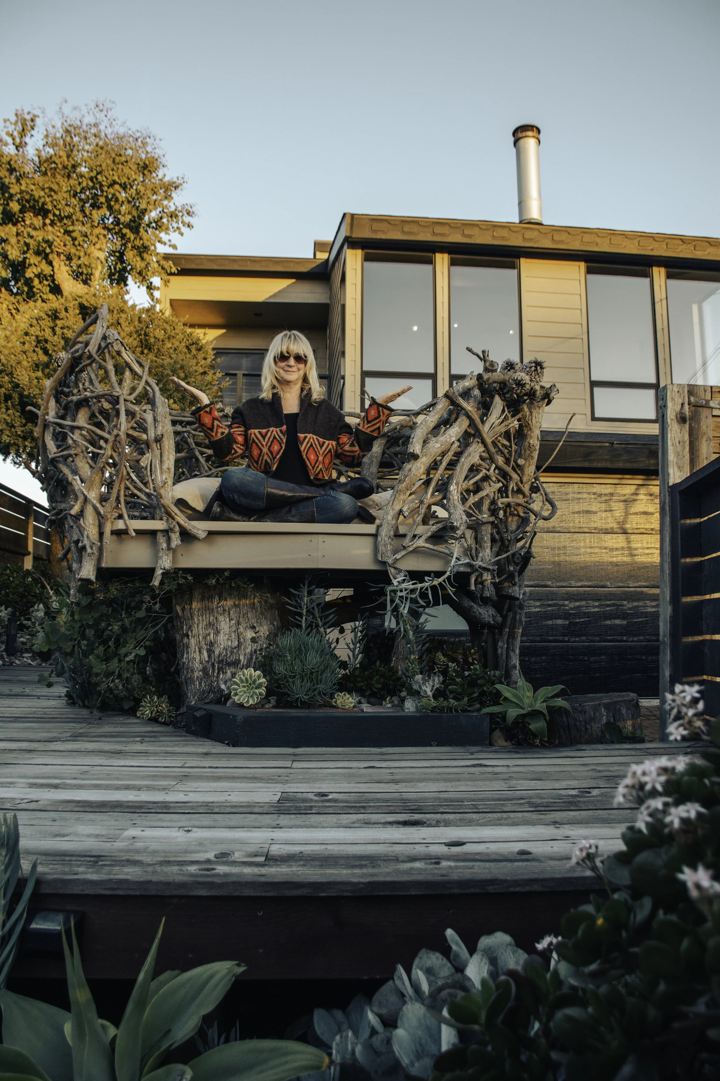Woman sitting cross-legged on a wooden deck decorated with plants, surrounded by trees and a modern house with large glass windows in the background, during sunset.