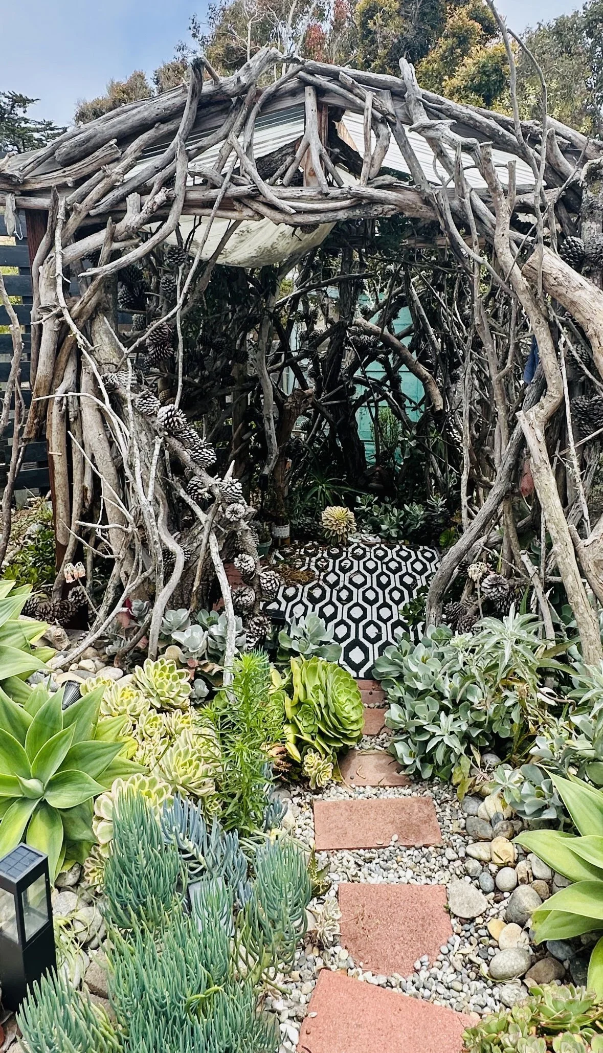 A garden path leading through a garden with various succulents and plants, with a small arbor made of driftwood branches and pinecones, and stepping stones on gravel.