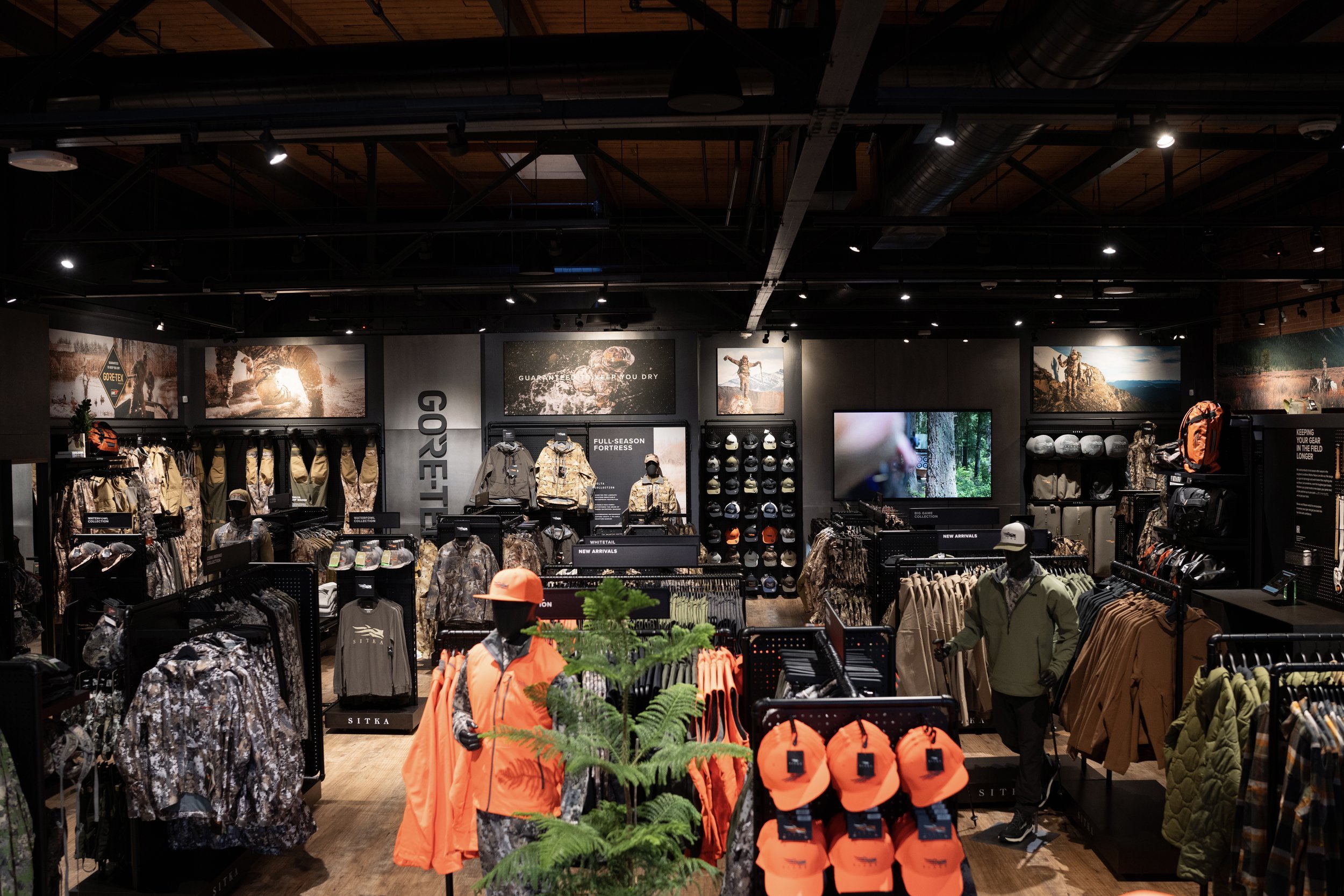 Interior of an outdoor gear store with clothing, hats, and helmets on display, including orange safety vests and caps, with posters of outdoor scenes on the wall and mannequins dressed in outdoor apparel.