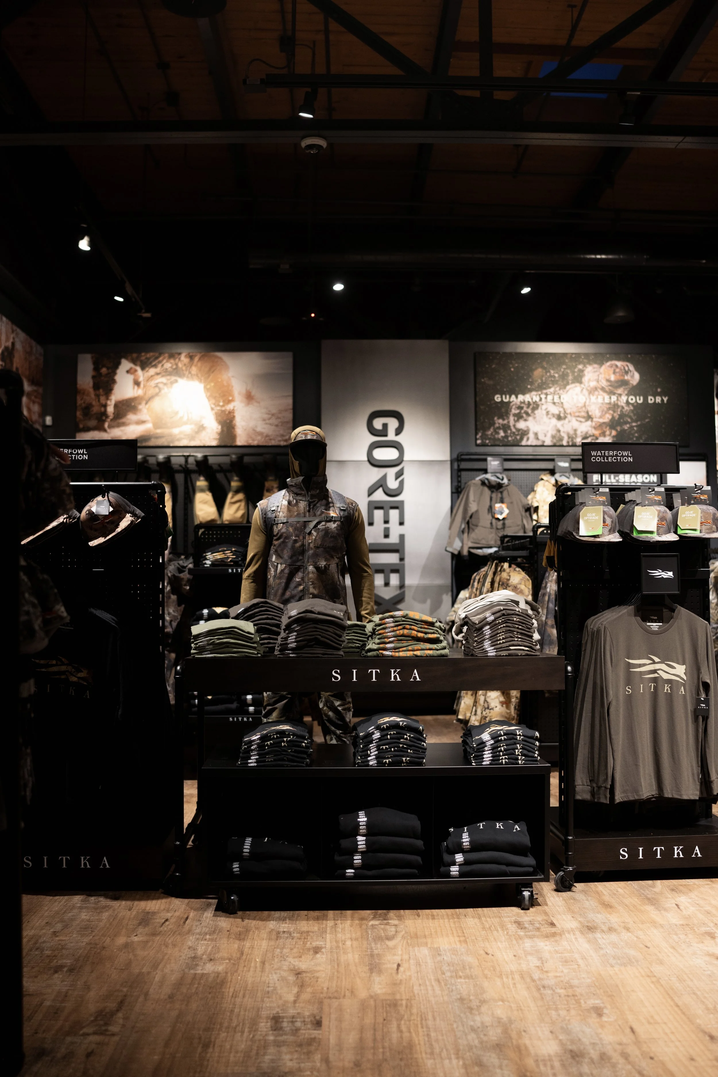 Indoor retail store displaying outdoor clothing and gear from the brand SITKA. Mannequins dressed in camouflage and outdoor apparel, with clothing racks and shelves filled with folded shirts and hats. Promotional posters with outdoor adventure imager