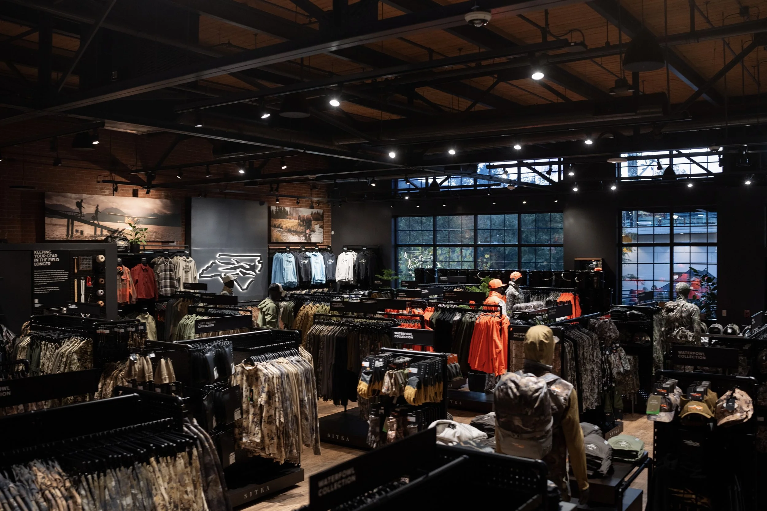 Interior of a retail store with clothing racks displaying jackets and shirts, large windows with natural light, brick walls, and a high ceiling with industrial lighting.