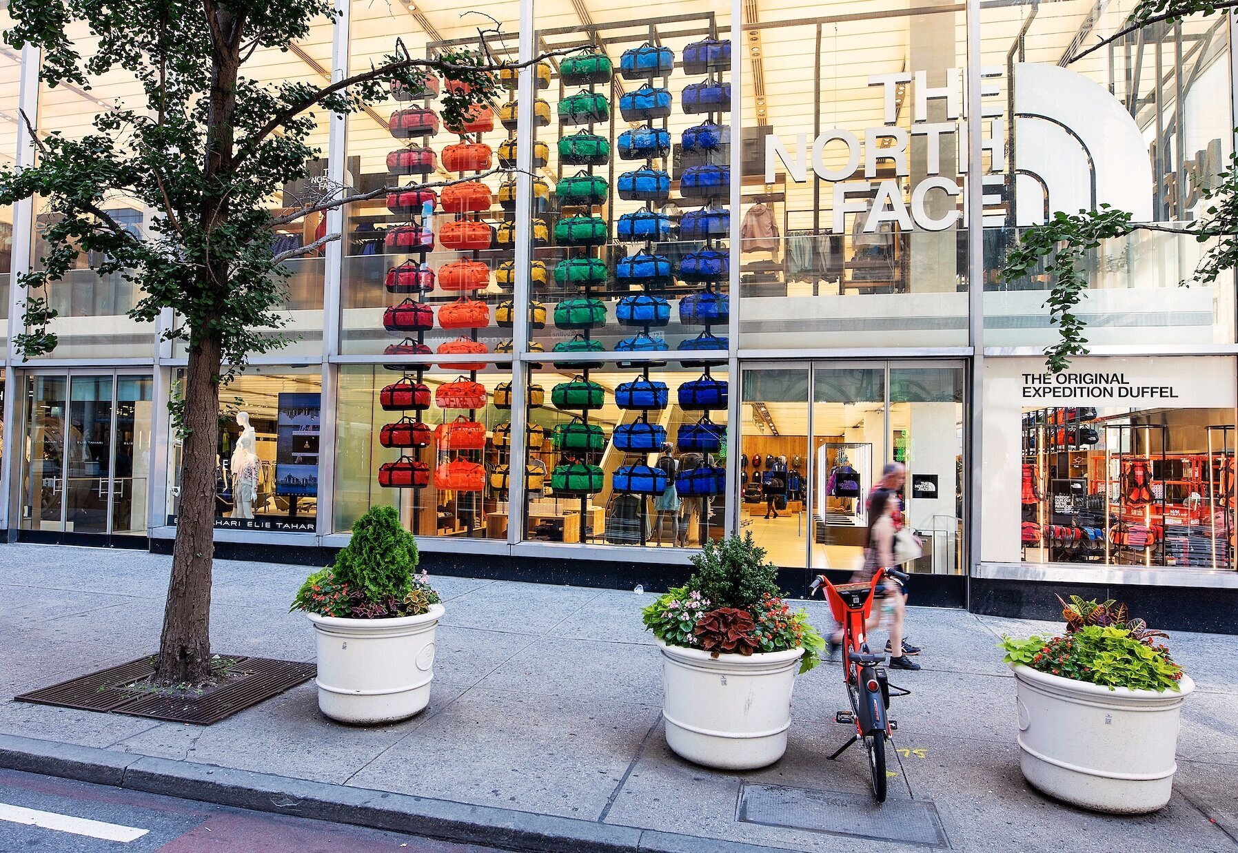 The storefront of The North Face store, displaying colorful backpacks arranged in a grid inside the glass window. Outside, there are large planters with flowers and trees, and a woman walking with a child on a scooter in front of the store.