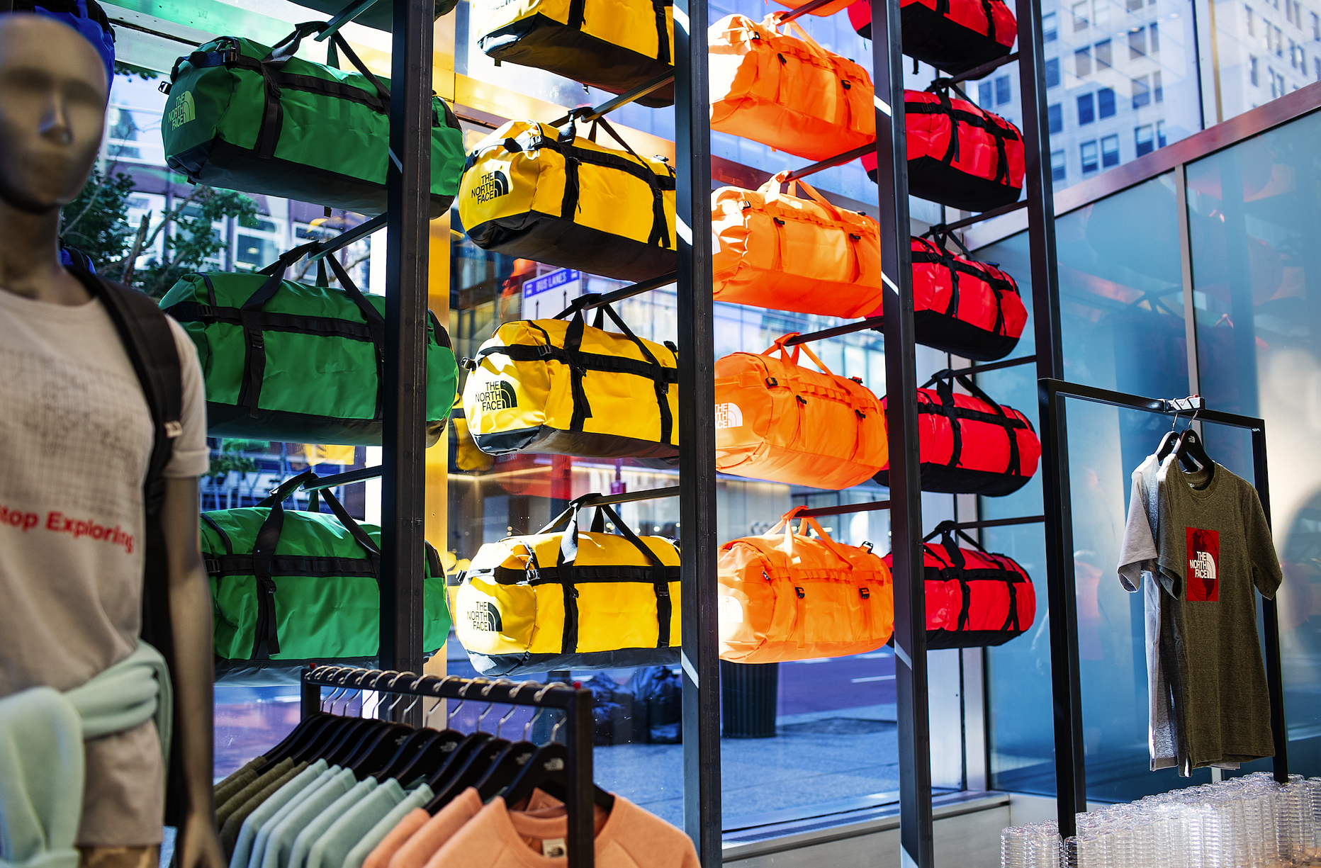Display of colorful duffle bags from The North Face on a wall shelf inside a retail store, with a mannequin torso in the foreground and clothing racks below.
