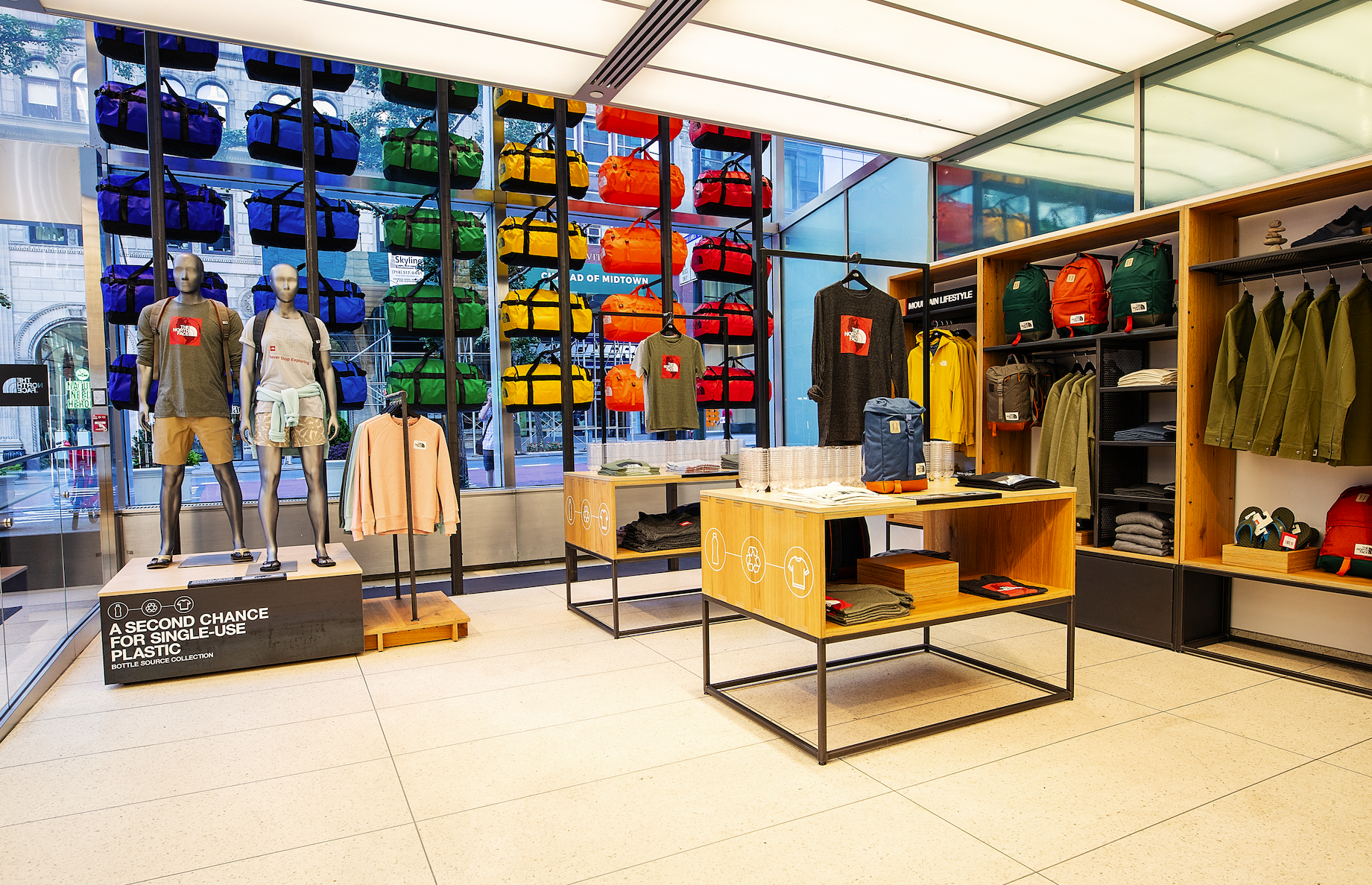 Clothing store display with mannequins dressed in casual wear, colorful bags on wall shelves, and wooden shelves with folded clothes in the background.