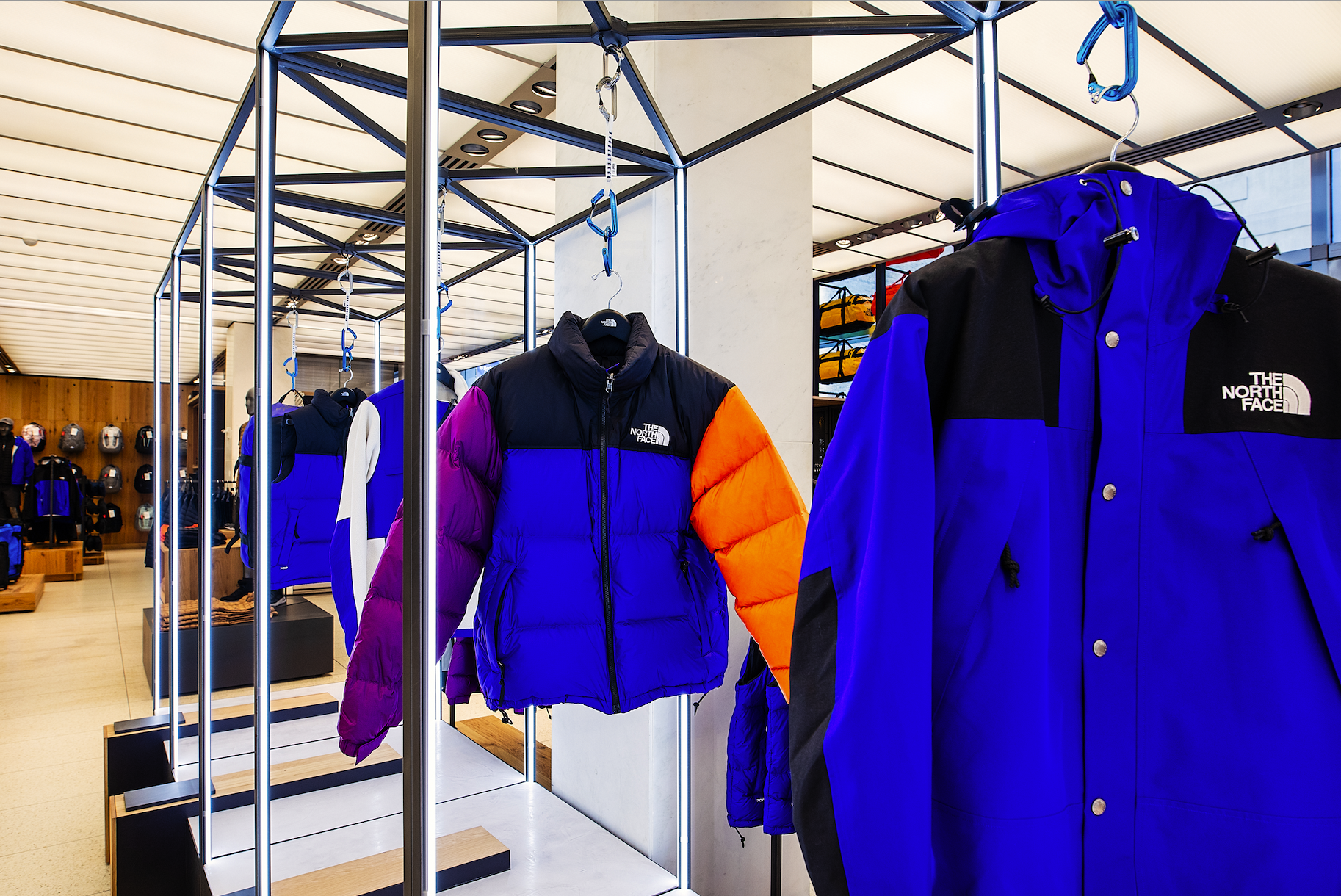 Display of blue, purple, black, and orange North Face jackets and outerwear on hangers in a retail store with shelves of backpacks in the background.