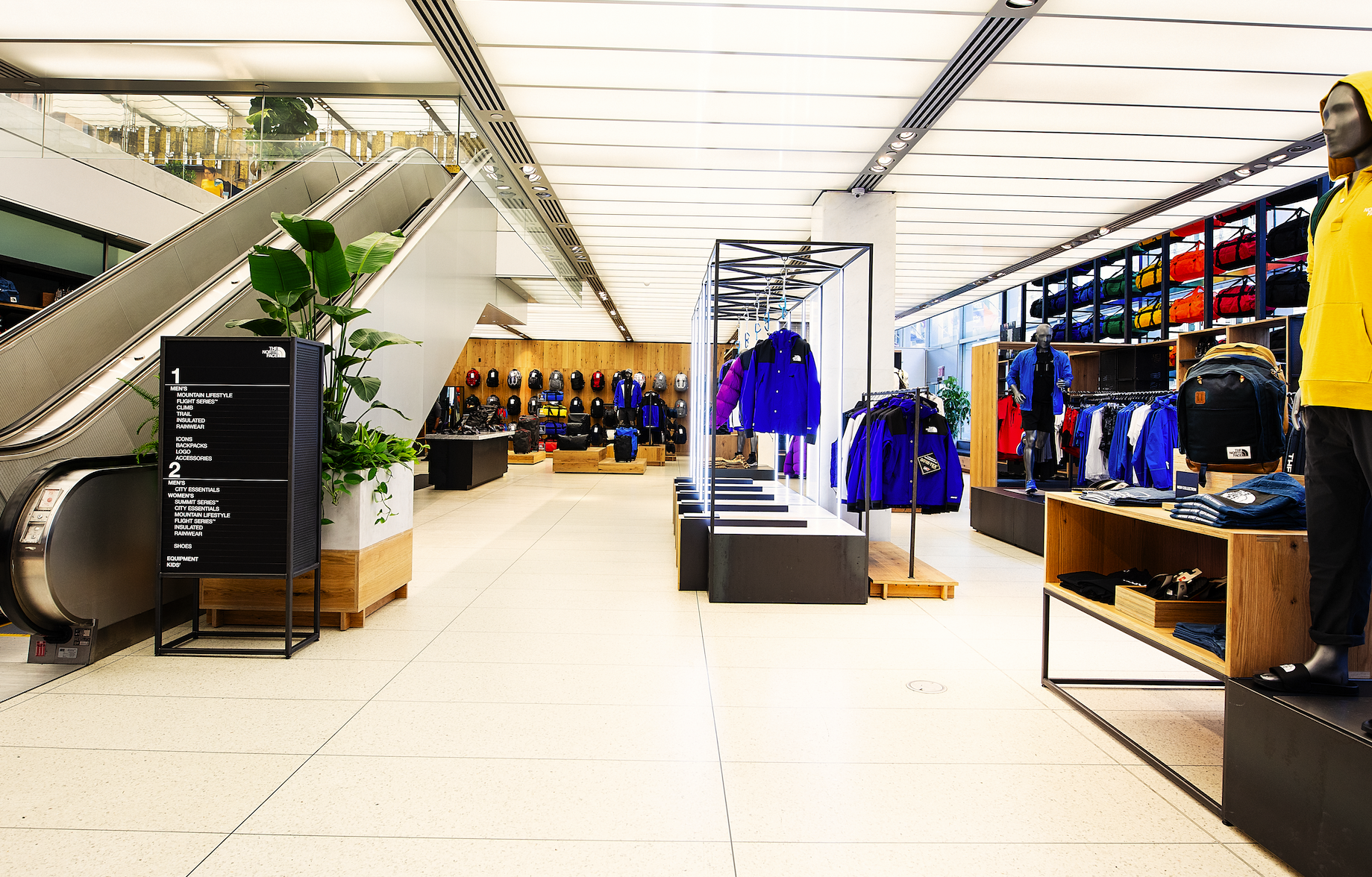 Inside a retail store with outdoor apparel and accessories, displaying jackets, backpacks, and helmets, near an escalator and a plant.