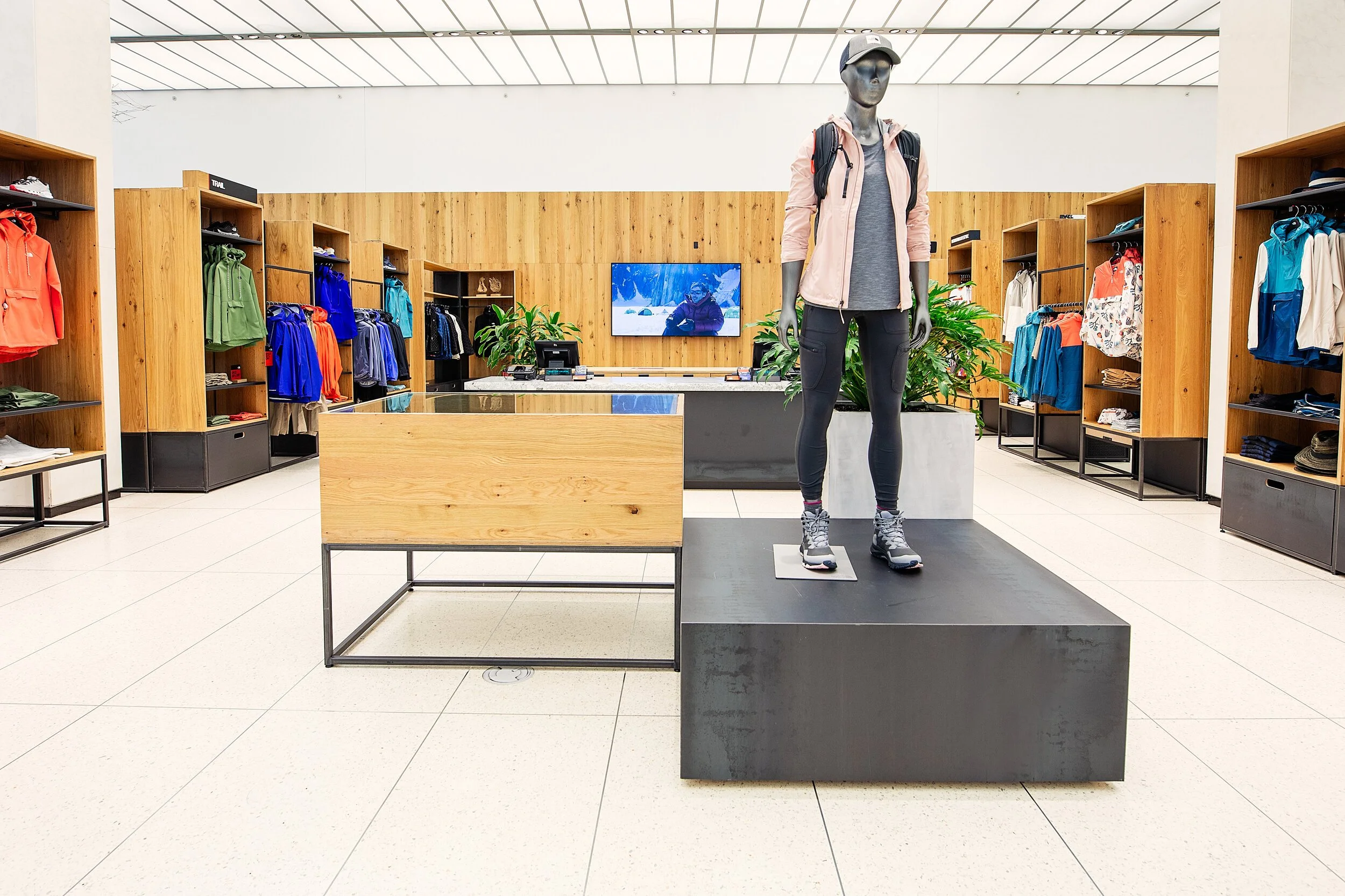 Clothing store with a mannequin dressed in outdoor gear, including a pink jacket, gray shirt, black leggings, hiking boots, and a gray cap. Shelves of colorful athletic wear surround the display.