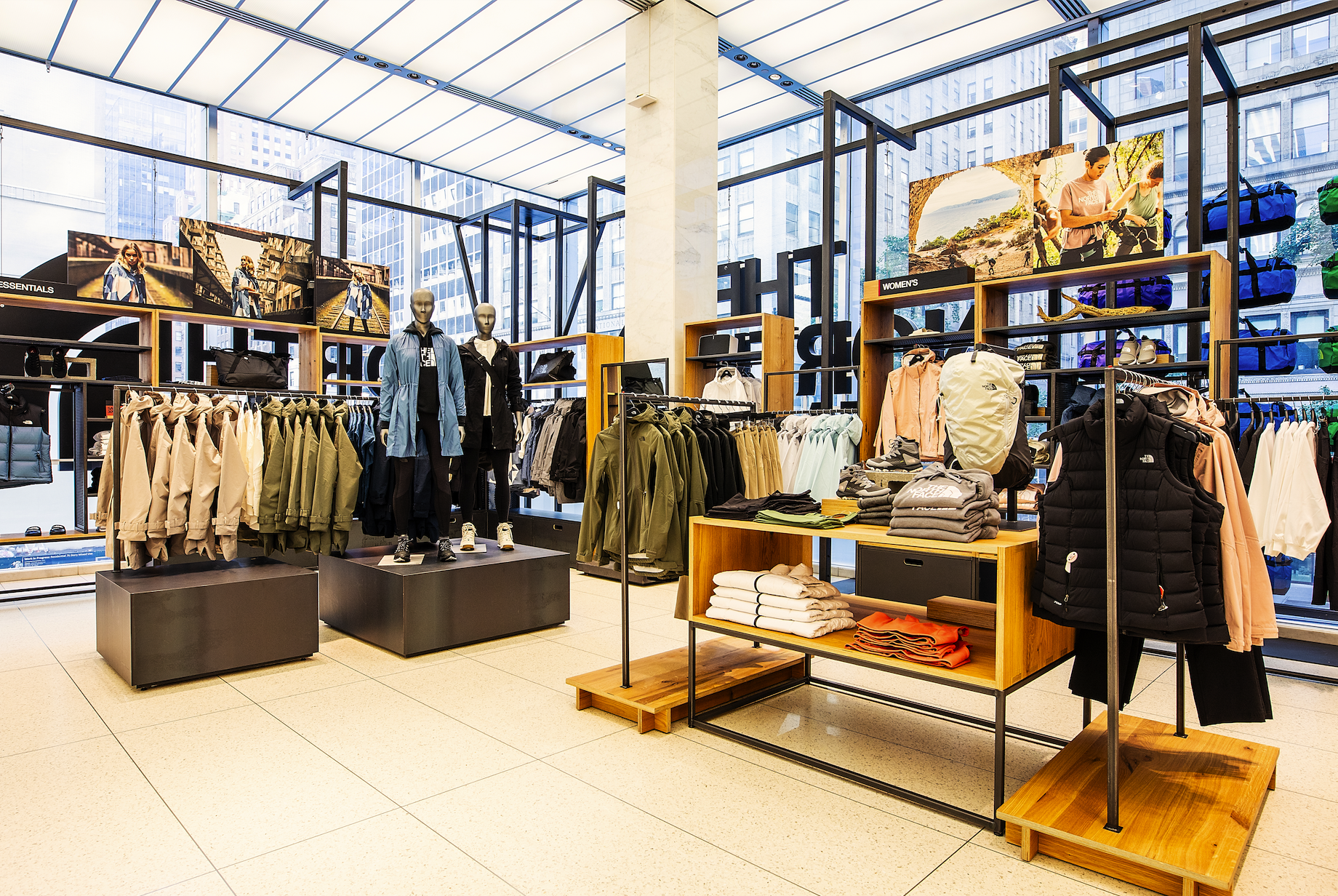 Inside a retail clothing store with windows showing city buildings, mannequins dressed in outdoor wear, and shelves with jackets, shirts, and accessories displayed.