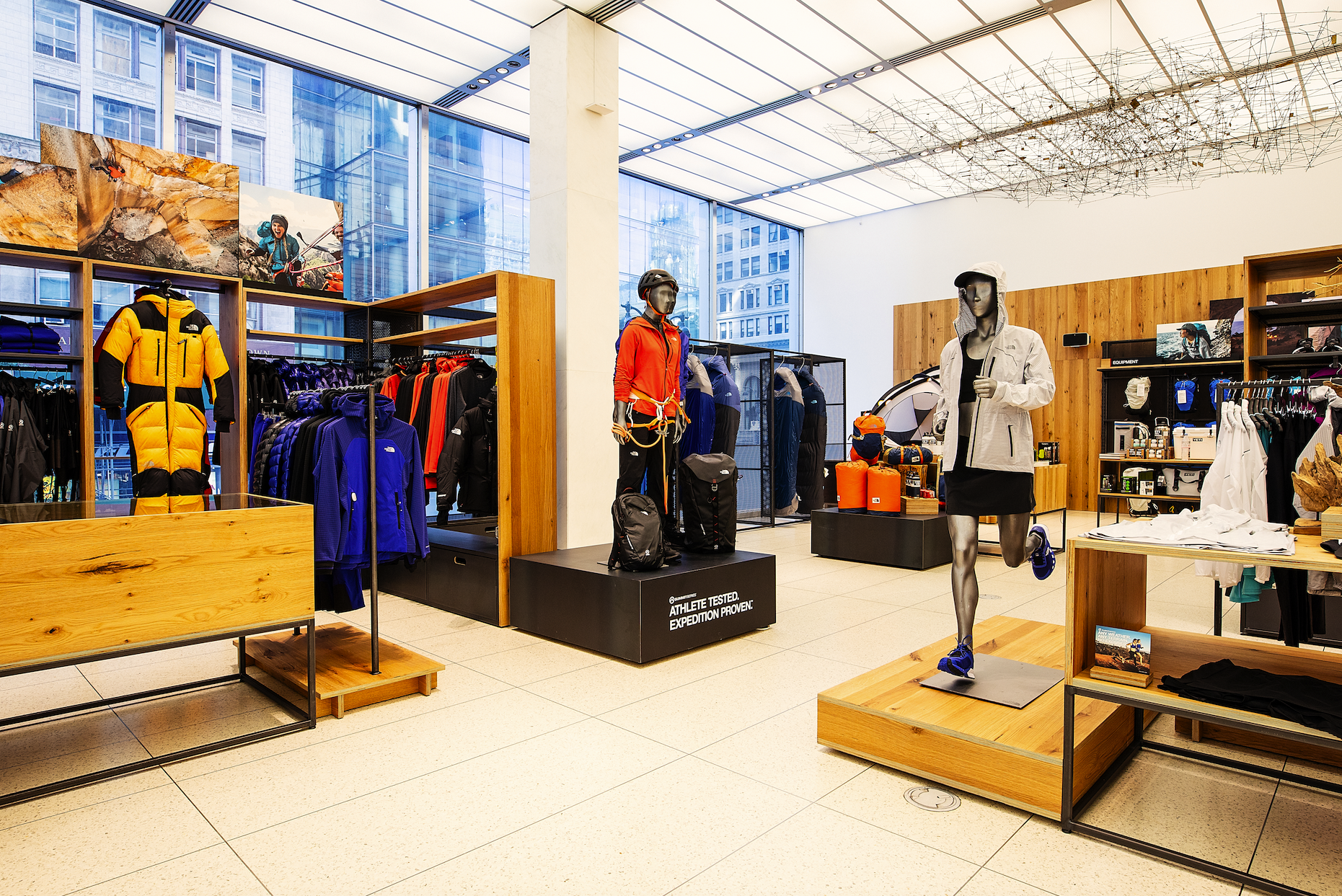 Athletic apparel and outdoor gear store with mannequins dressed in outdoor clothing, backpacks, and climbing equipment, in a modern retail space with large windows and wooden fixtures.