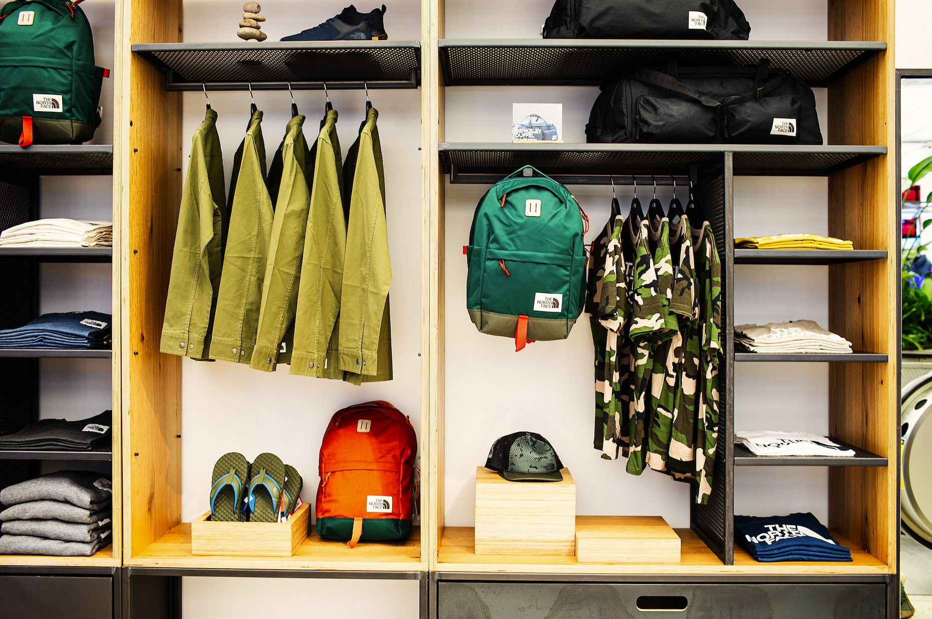 Custom closet with green jackets, camouflage shirts, backpacks, black duffel bags, folded clothes, slippers, and hats on shelves and hangers.
