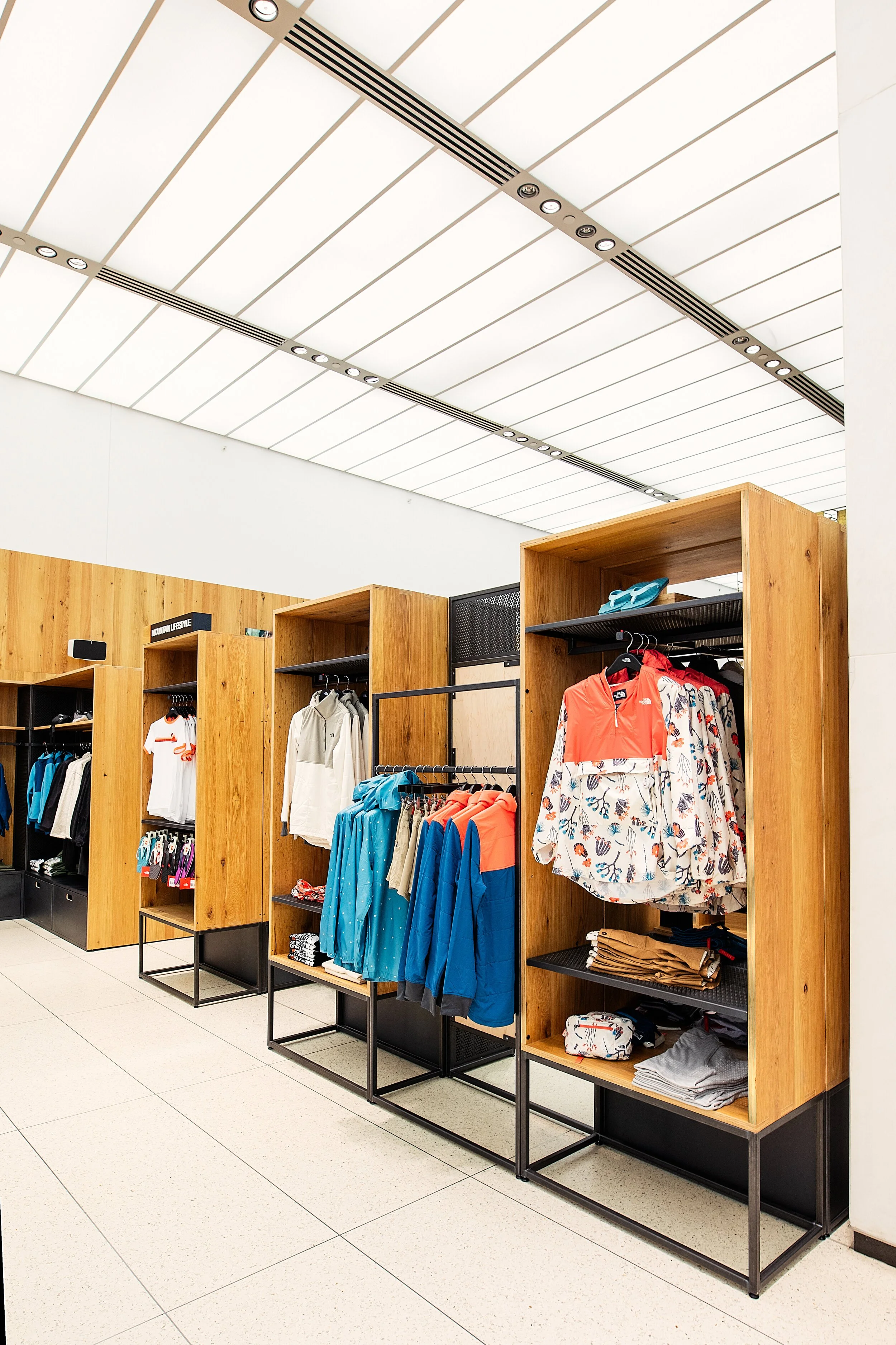 Clothing displays in a retail store with wooden shelves, showcasing sportswear, jackets, shirts, and accessories. The ceiling has modern lighting and air vents.