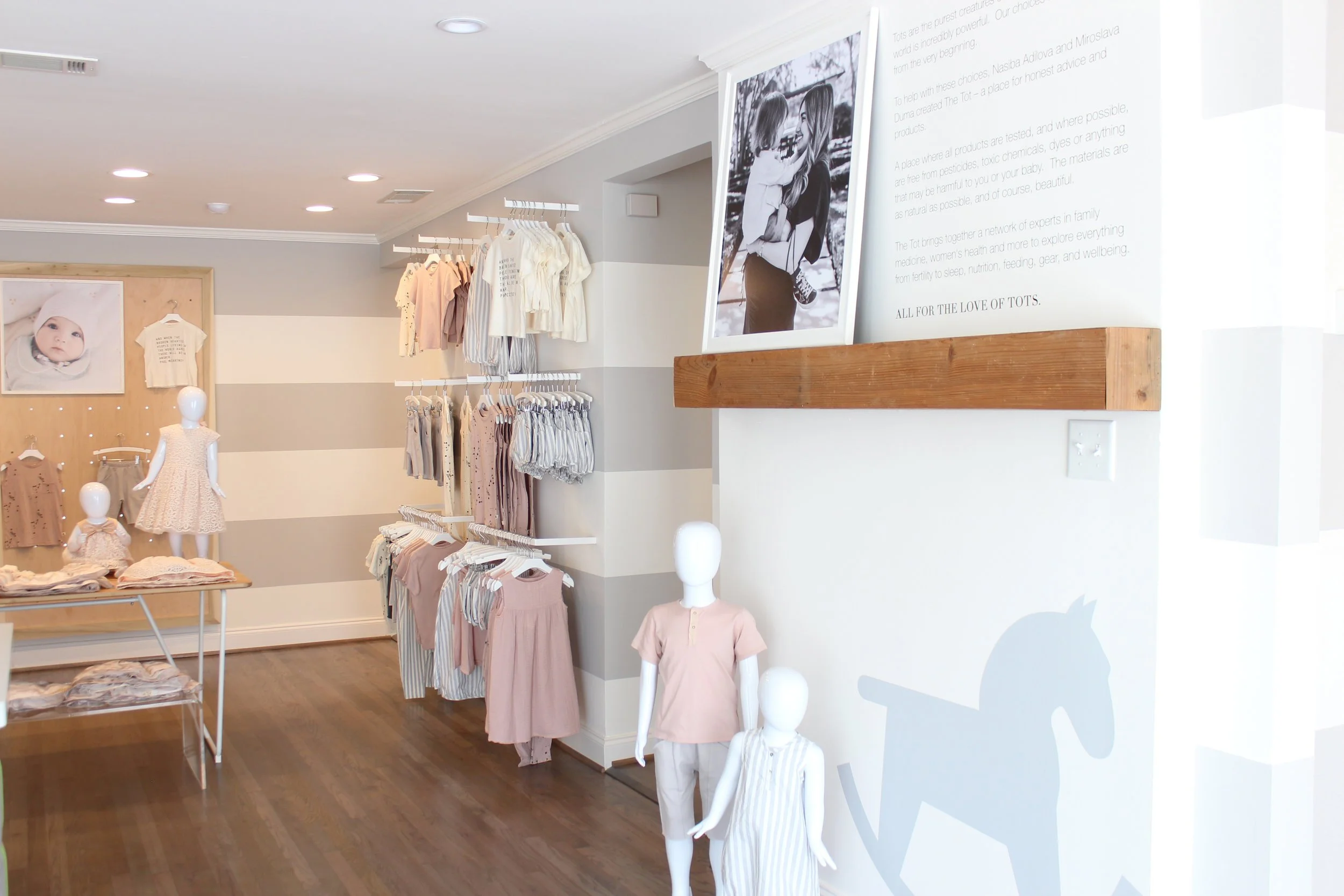 Children's clothing display in a boutique store with mannequins wearing pink and striped dresses, and racks of pastel-colored shirts and pants. A large black and white photo on the wall showcases a parent and child, and a wooden feature wall with fra