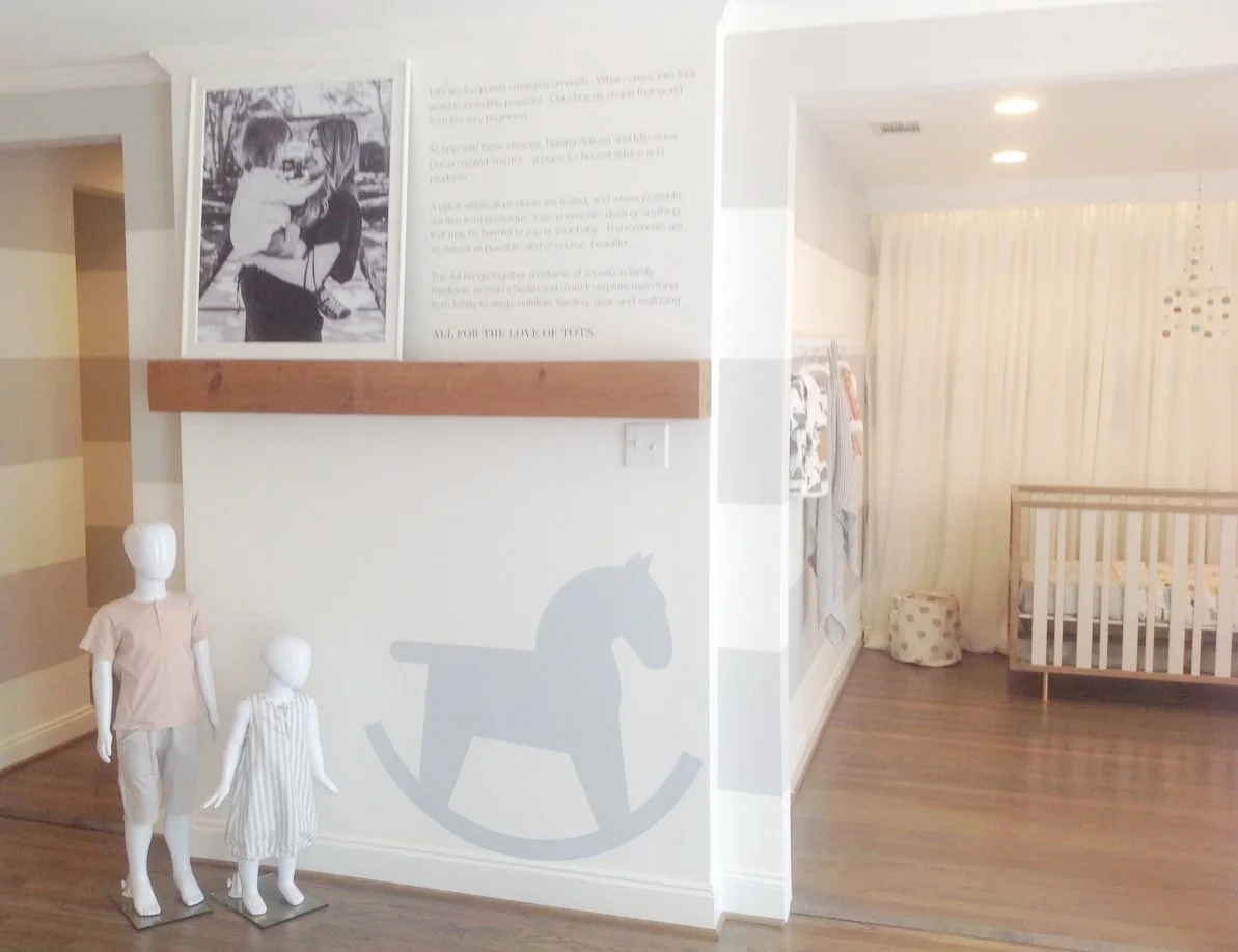 Interior of a nursery or baby's room with a white wall decorated with a gray rocking horse silhouette, two child mannequins dressed in children's clothing, a framed black and white photo of children, and a wall with printed text. The room has hardwood flooring, a crib, and a cream-colored curtain.
