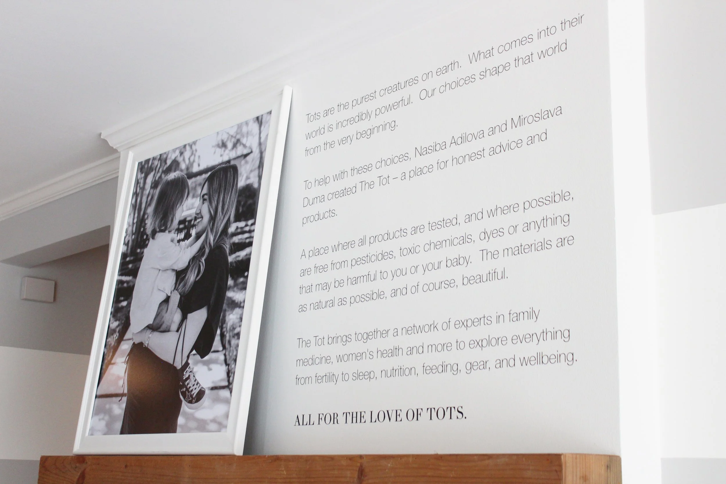 A wall with a framed black and white photograph of two young girls smiling and hugging, and a large block of text next to it discussing topics related to tots, children, and family, in an indoor setting with a white wall and crown molding.