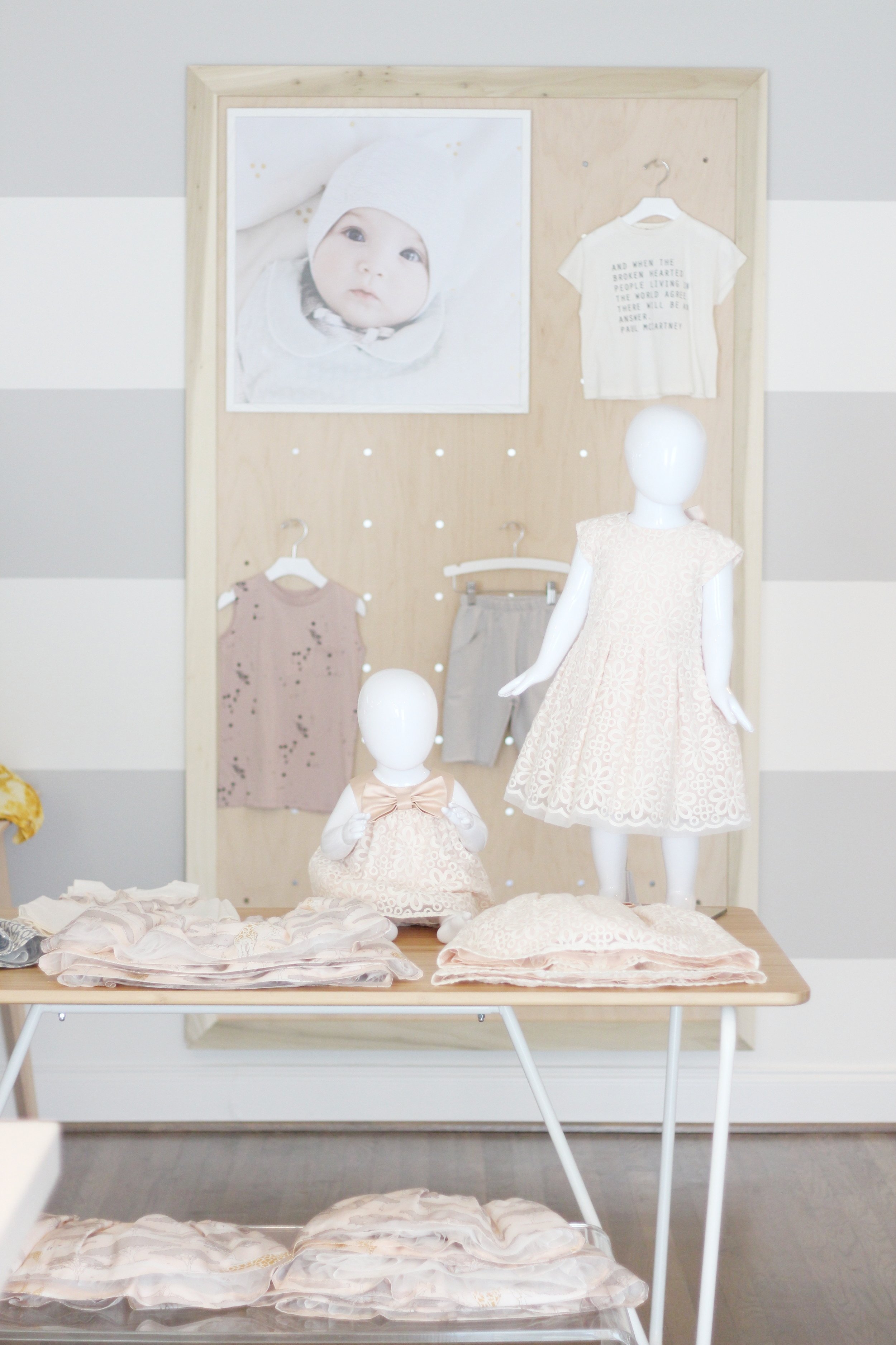 Display of children's clothing and accessories on mannequins and a table, with a framed photograph of a baby and a small white t-shirt with text, in a store or showroom setting.