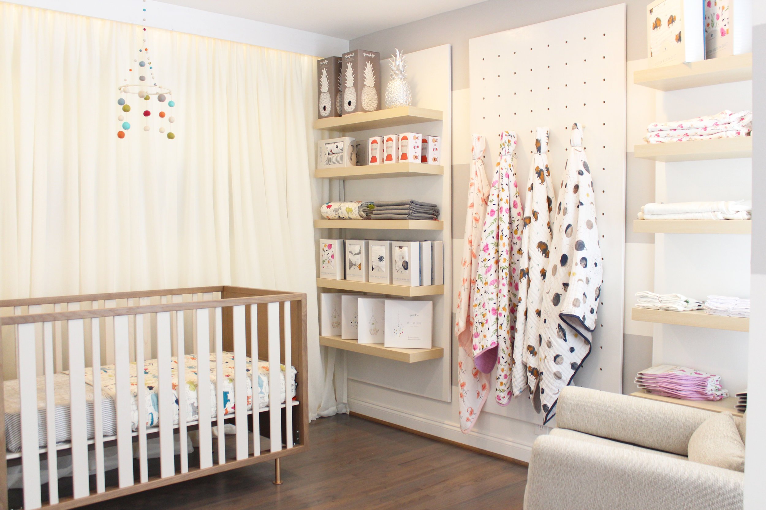 Nursery room with a wooden crib, cream curtains, and shelves filled with folded blankets and boxes, some decorated with baby-themed patterns and images.