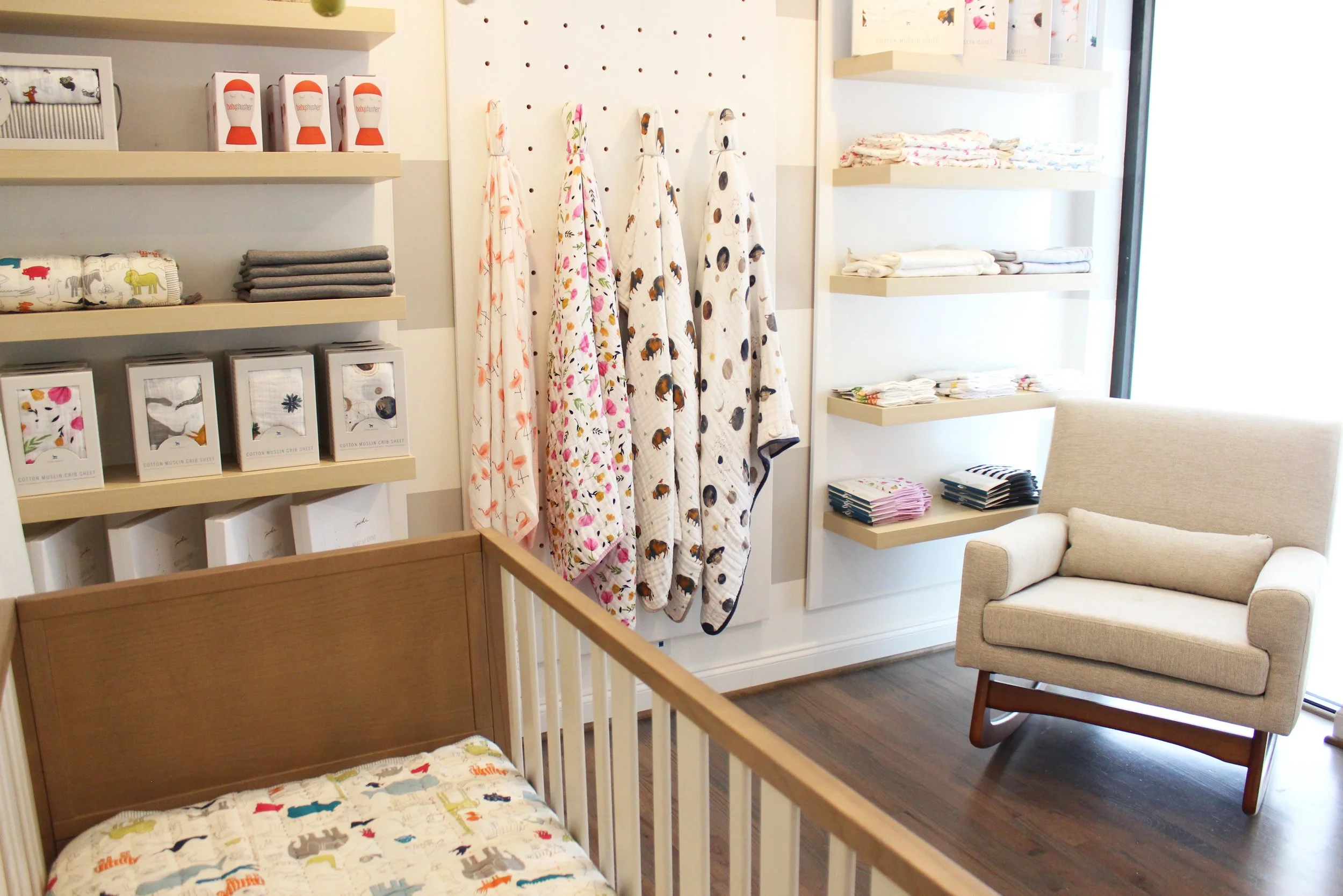 A display of baby products in a retail store, including patterned blankets hanging on a pegboard, neatly folded clothing on shelves, and a crib with a jungle-themed blanket, with a comfortable armchair nearby.