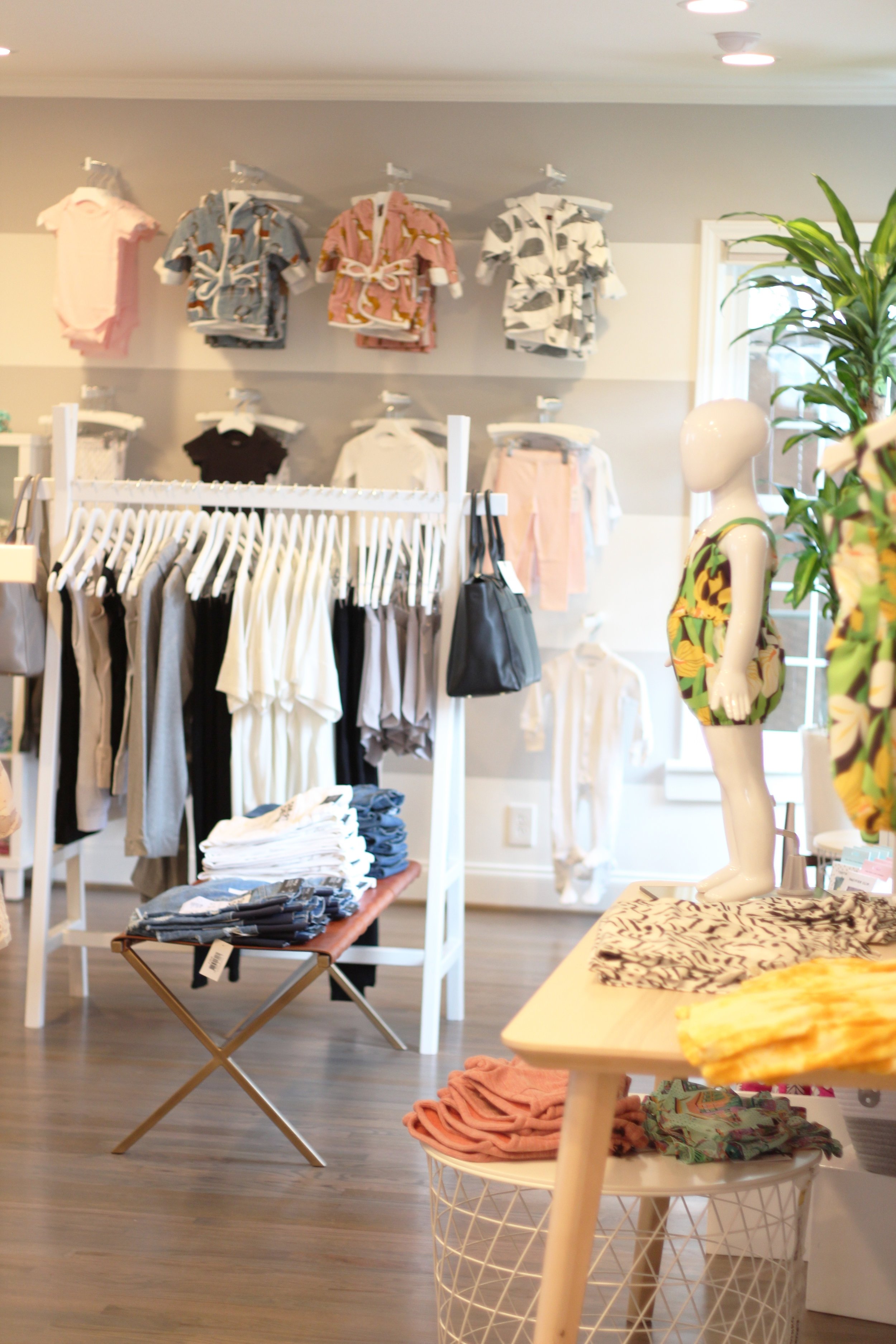 Clothing display in a boutique, with folded clothes on a table, hanging garments on racks, and mannequins dressed in colorful outfits.