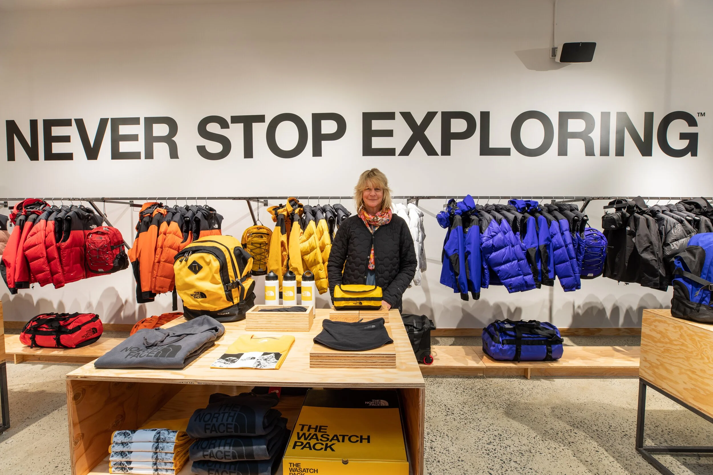 A woman standing behind a display table with North Face outdoor gear, including jackets, backpacks, water bottles, and folded clothing, in front of a wall with the words 'Never Stop Exploring' in large black letters.
