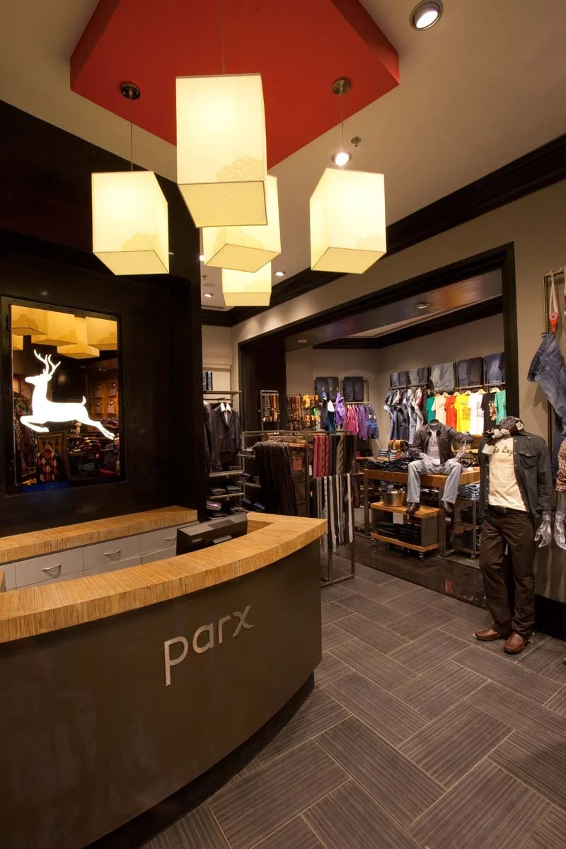 Interior of a clothing store with a wooden checkout counter labeled 'parx,' a chandelier with multiple rectangular lights hanging from a red ceiling, and mannequins displaying casual clothes with shelves and racks of apparel in the background.