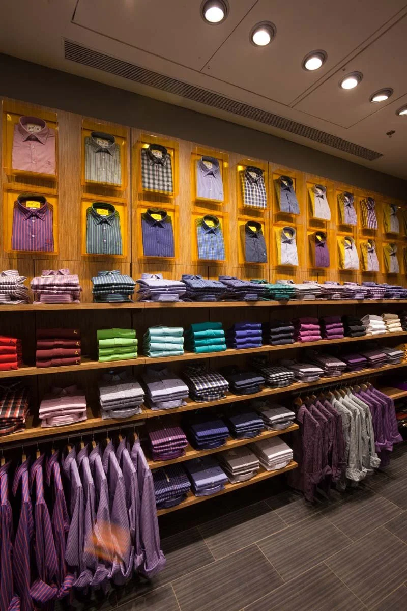 Display of neatly folded and hanging men's dress shirts in various colors and patterns on wooden shelves in a retail store.