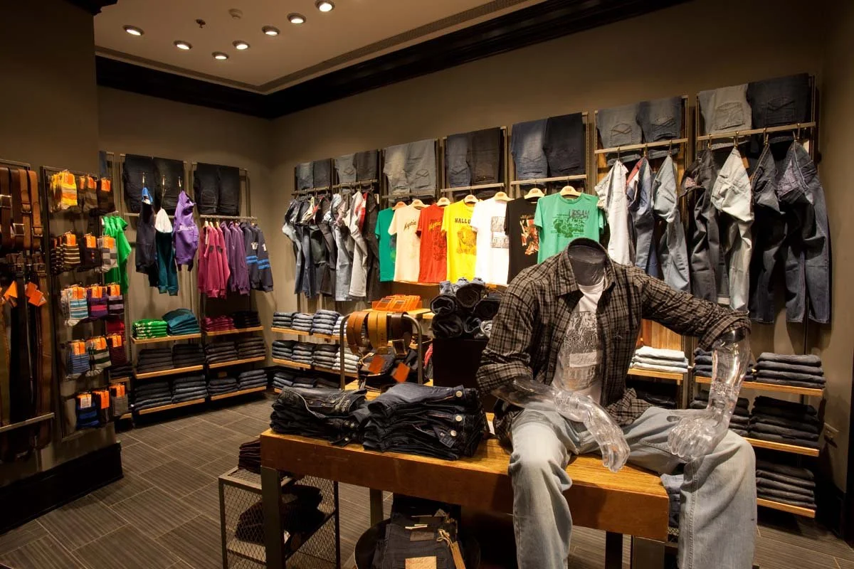 Clothing store display with various pants, shirts, and accessories. A mannequin dressed in casual clothes sits on a wooden bench.