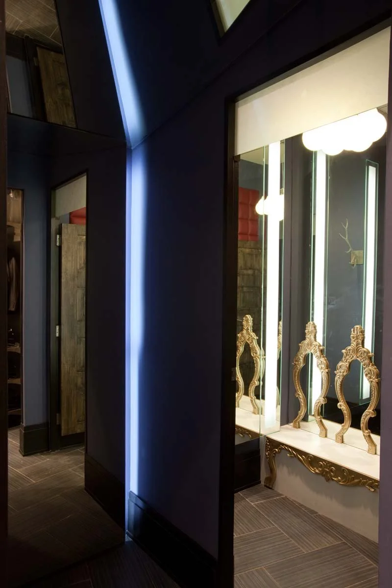 Interior of a modern room with dark walls and wooden furniture, featuring illuminated decorative mirrors with ornate gold frames, and a light fixture with white, cloud-like bulbs.