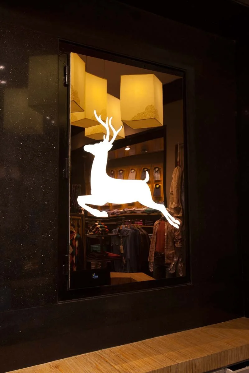 A glass window with a white silhouette of a leaping deer. Inside the background includes folded clothing and hanging shirts, with warm ceiling lights and a decorative hanging lamp.