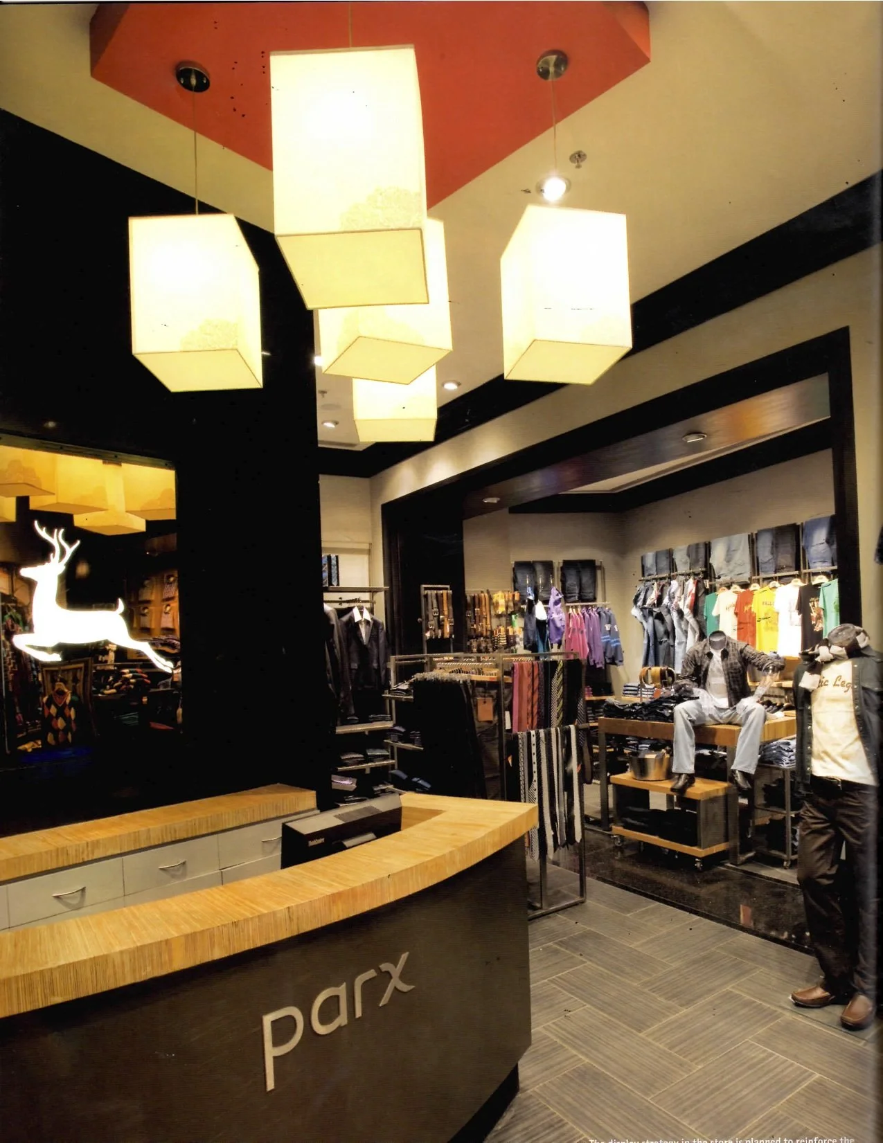 Inside a retail clothing store with a wooden counter in the foreground and clothing racks with mannequins in the background, illuminated by square hanging ceiling lights.