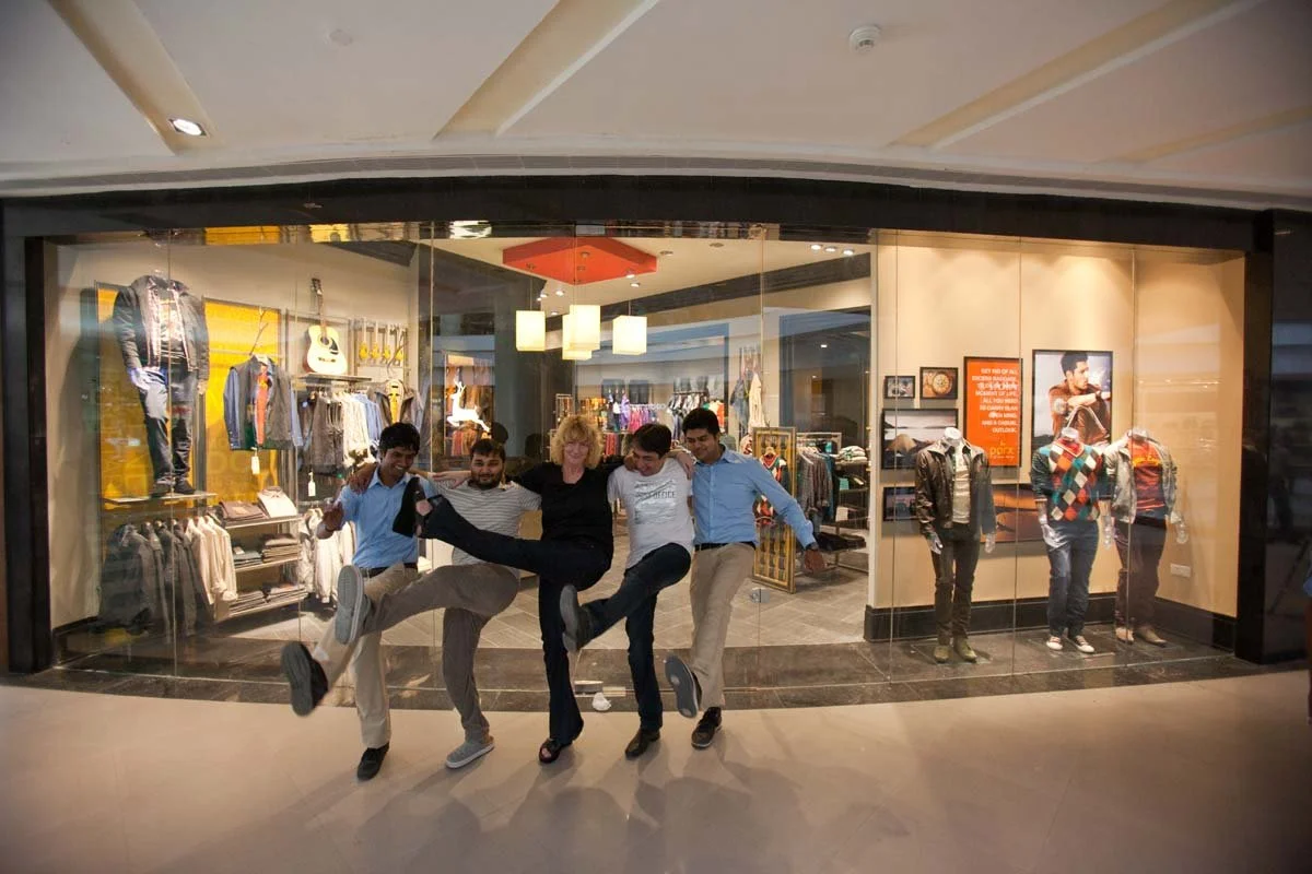 Four young men standing outside a clothing store, smiling, with their legs raised and arms around each other's shoulders, posing playfully in front of the store window.