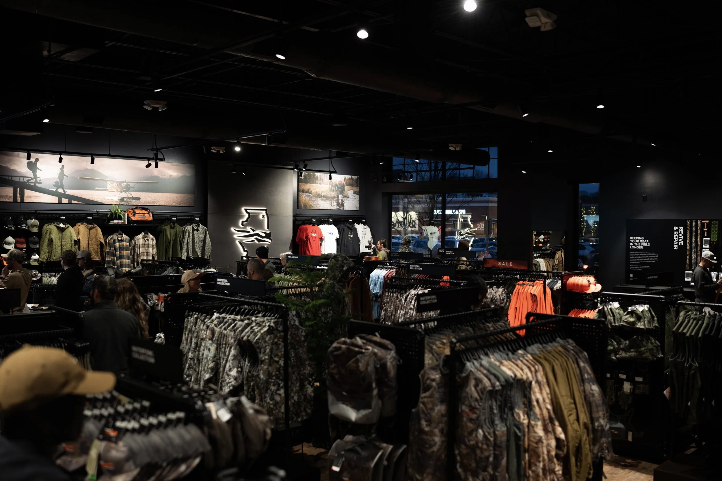 Inside of a retail store selling outdoor and adventure clothing and gear, with racks of jackets, shirts, hats, and accessories, and customers shopping.