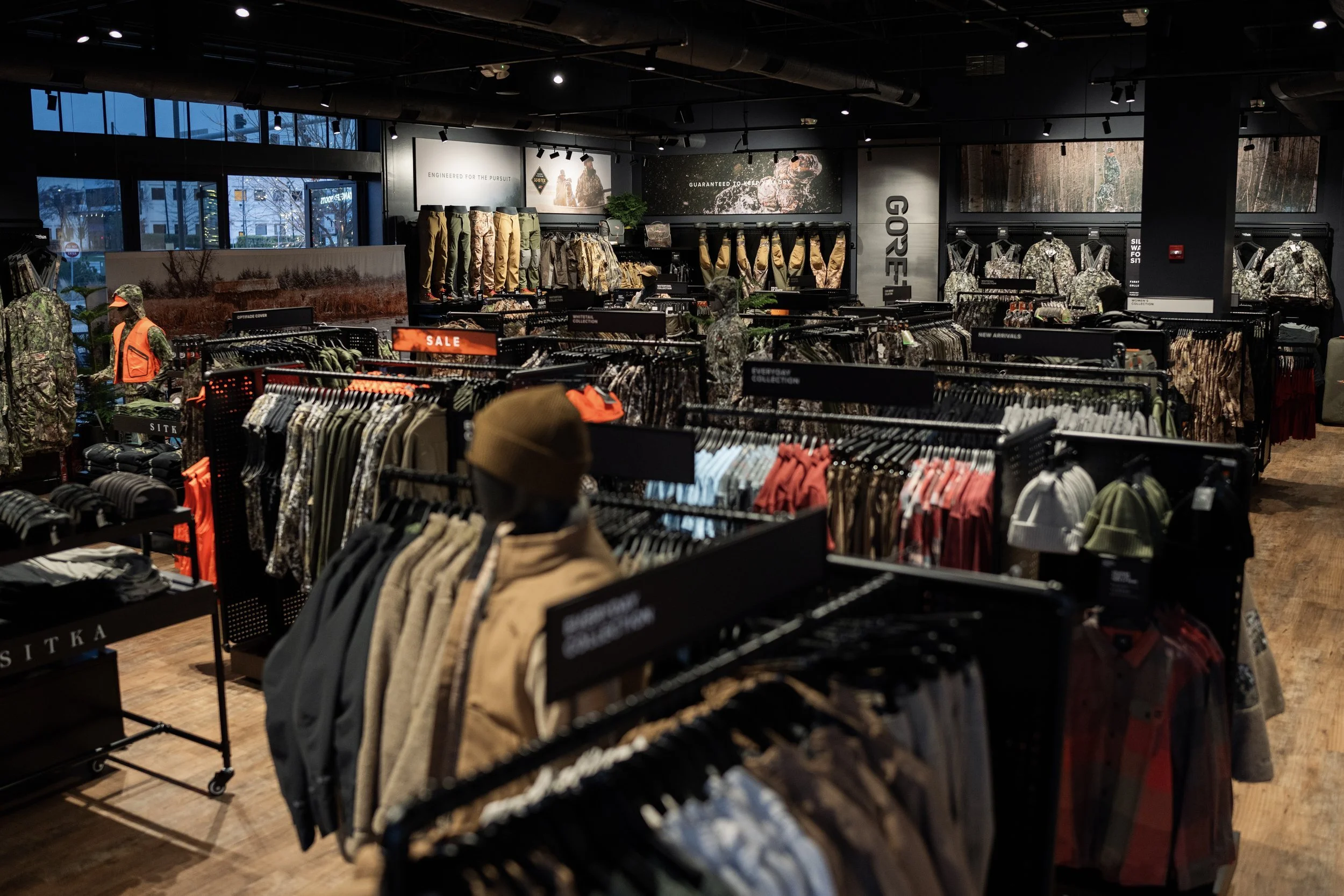 Indoor retail store selling outdoor and military-style clothing and gear, with racks of clothing, mannequins in camouflage, and signs indicating sale and various collections.